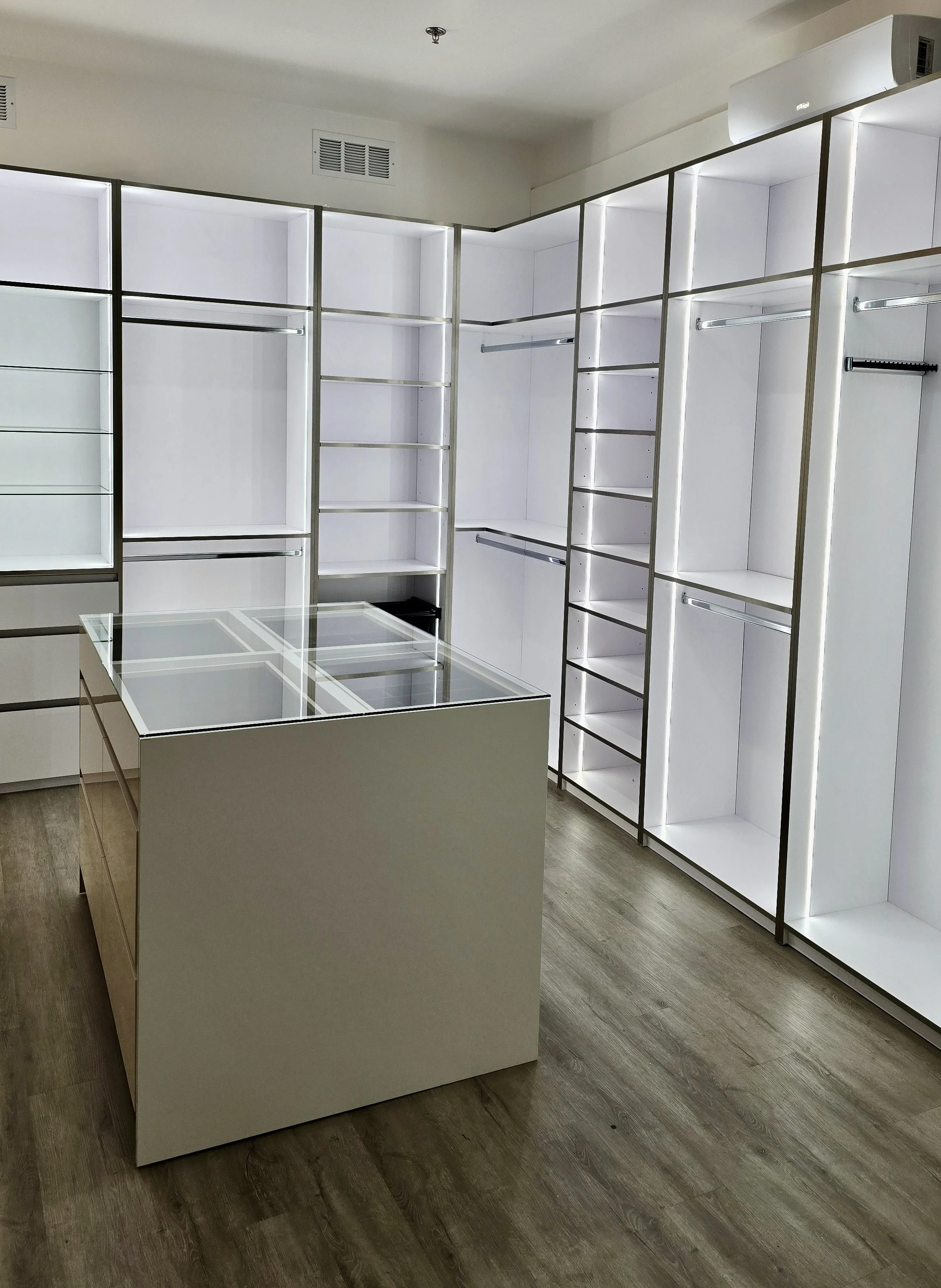 Spacious, modern walk-in closet with white shelving and hanging rods, illuminated by LED lights. A central island with glass top is surrounded by open storage spaces. The floor is hardwood.