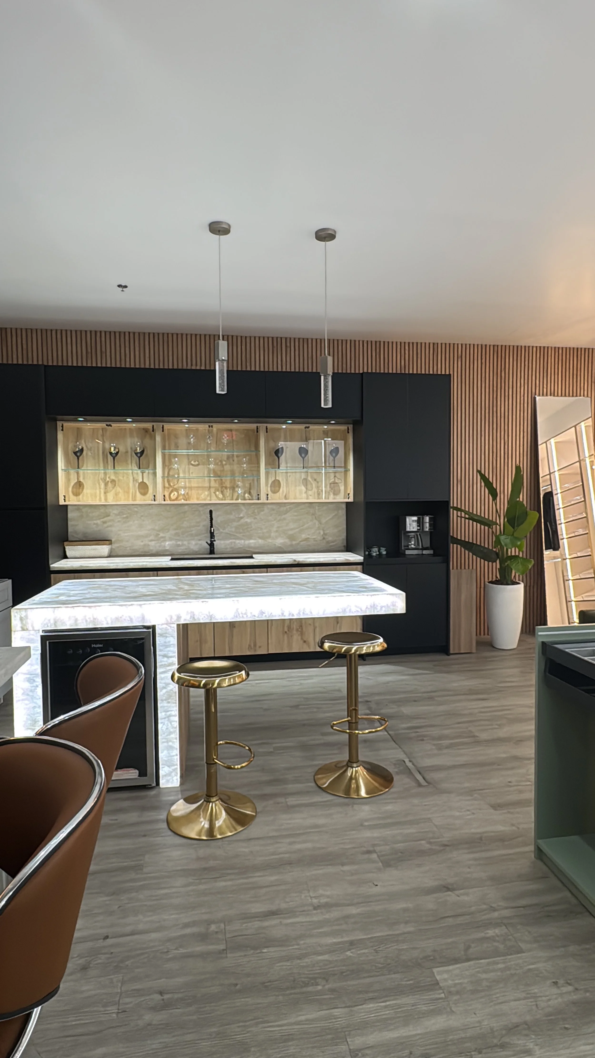 Modern kitchen with illuminated white translucent quartzite island, black and wooden cabinetry, gold bar stools, and a large potted plant.