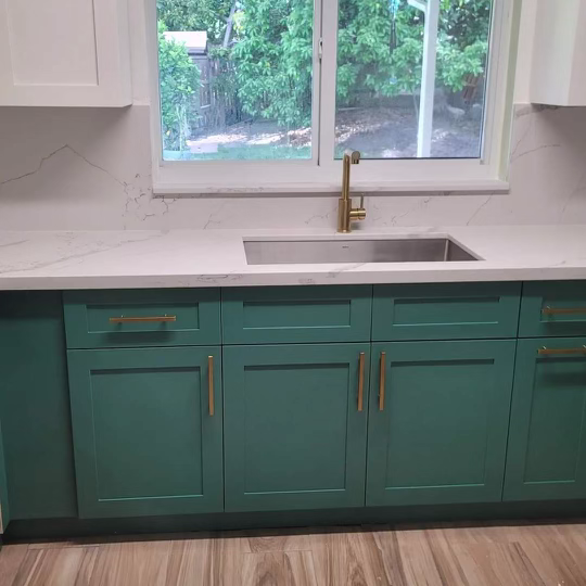 Modern kitchen with green teal cabinets and gold handles, white quartz countertop, undermount sink, gold faucet, wooden floor, and window overlooking garden.
