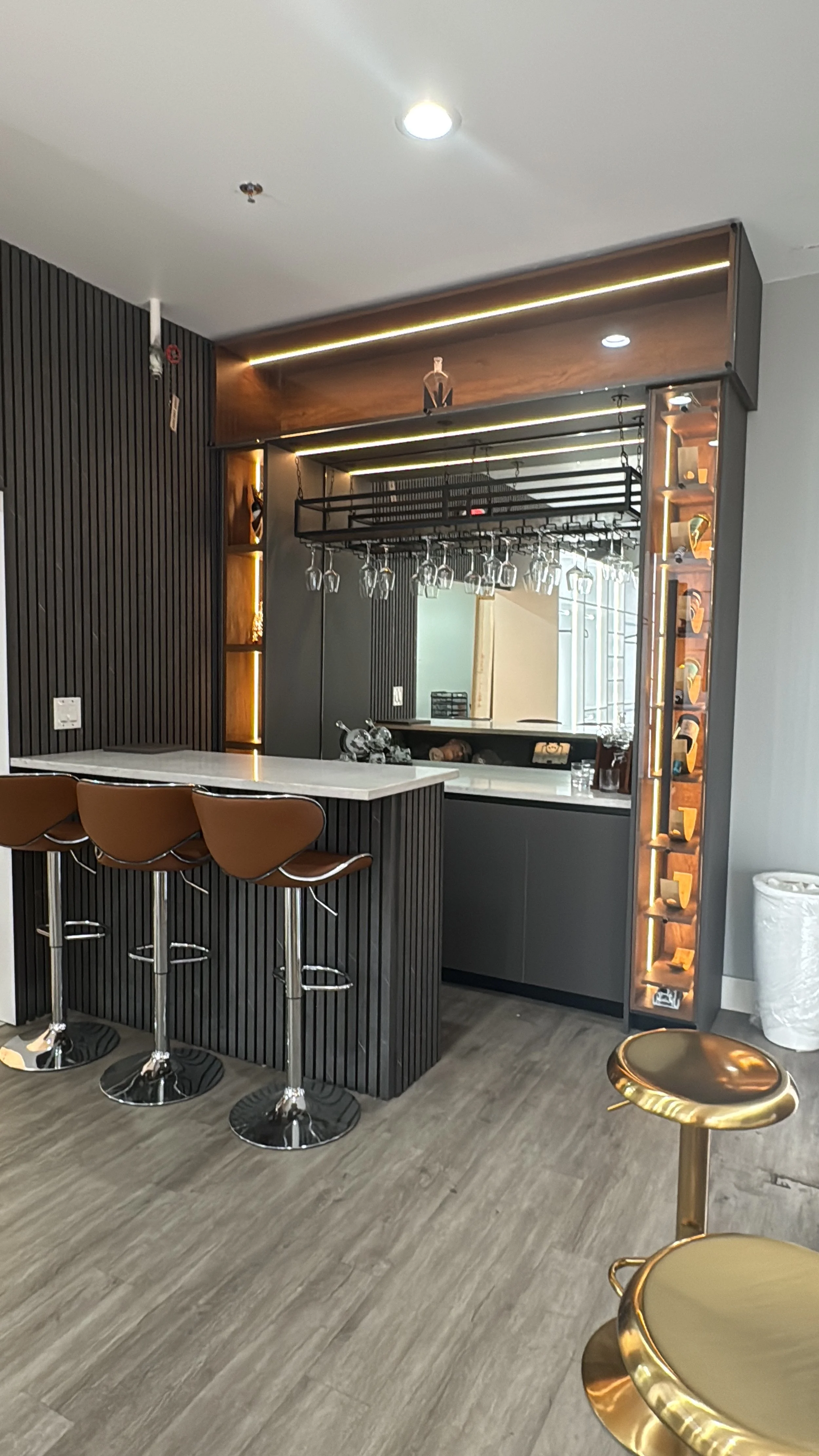 Modern home bar area with a white granite counter, brown bar stools, wine glasses hanging upside down, a wine rack with bottles, and decorative lighting.