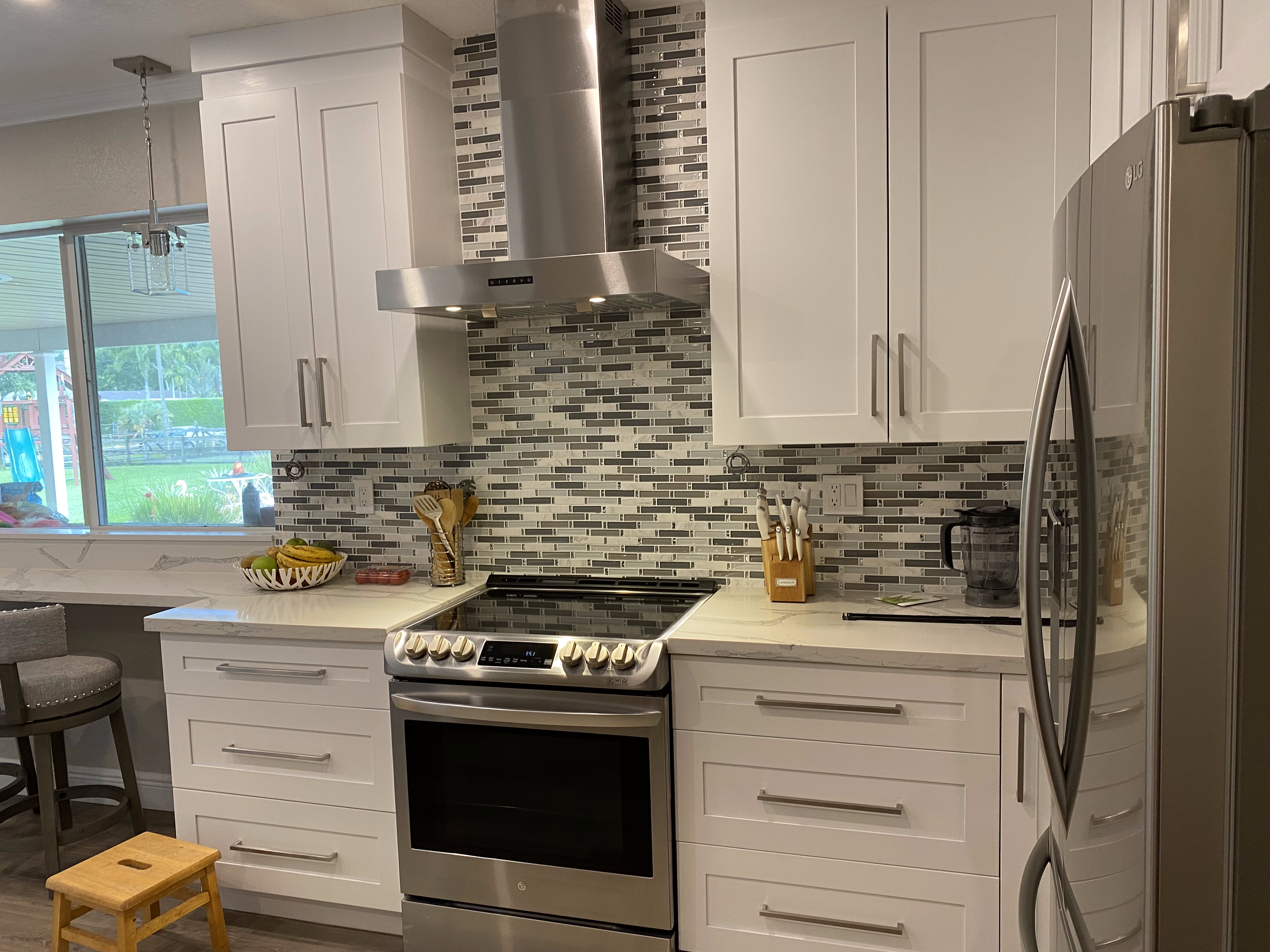 Modern kitchen with white cabinets, a stainless steel range, a mosaic tile backsplash, and a stainless steel refrigerator. A bowl of fruit and utensils are on the countertop.