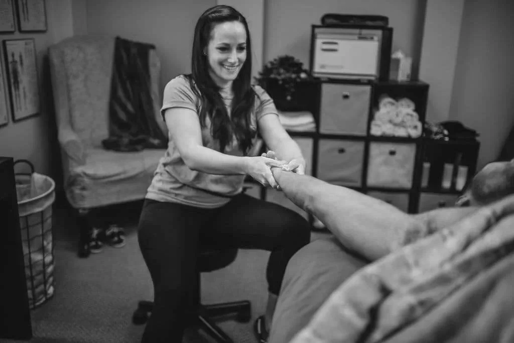 A woman smiling and pulling a man's hand toward her while sitting on a chair in a room with a bed, a laundry basket, and a cabinet with a computer monitor.