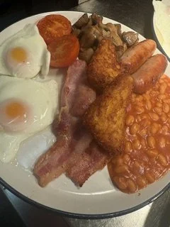 Full breakfast plate with fried eggs, tomato slices, fried chicken, bacon, sausage links, baked beans, and a hash brown.
