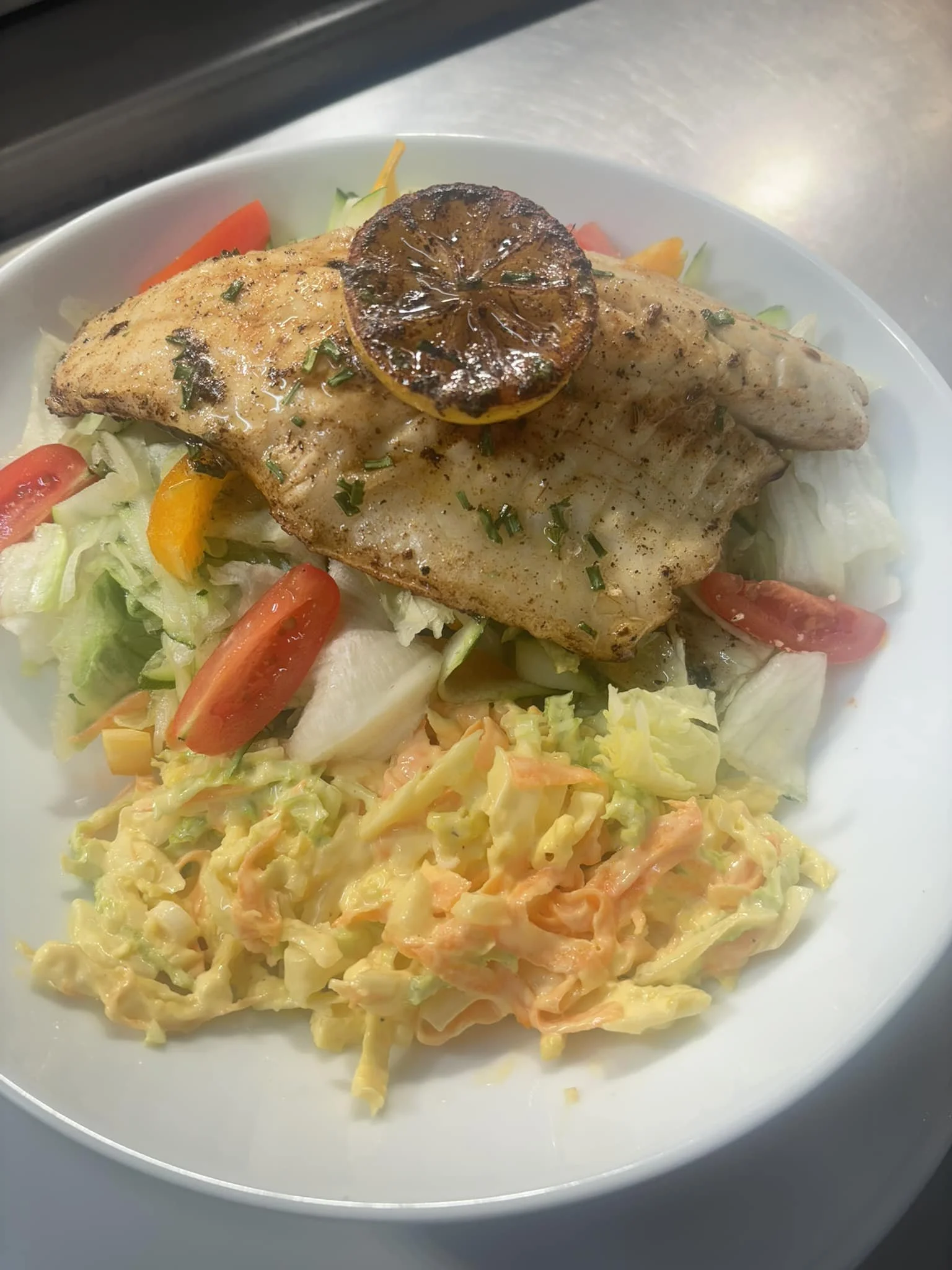 A plate with grilled chicken topped with a lemon slice, served on a bed of mixed vegetables, including lettuce, cherry tomatoes, and shredded carrots, along with a side of macaroni and cheese.