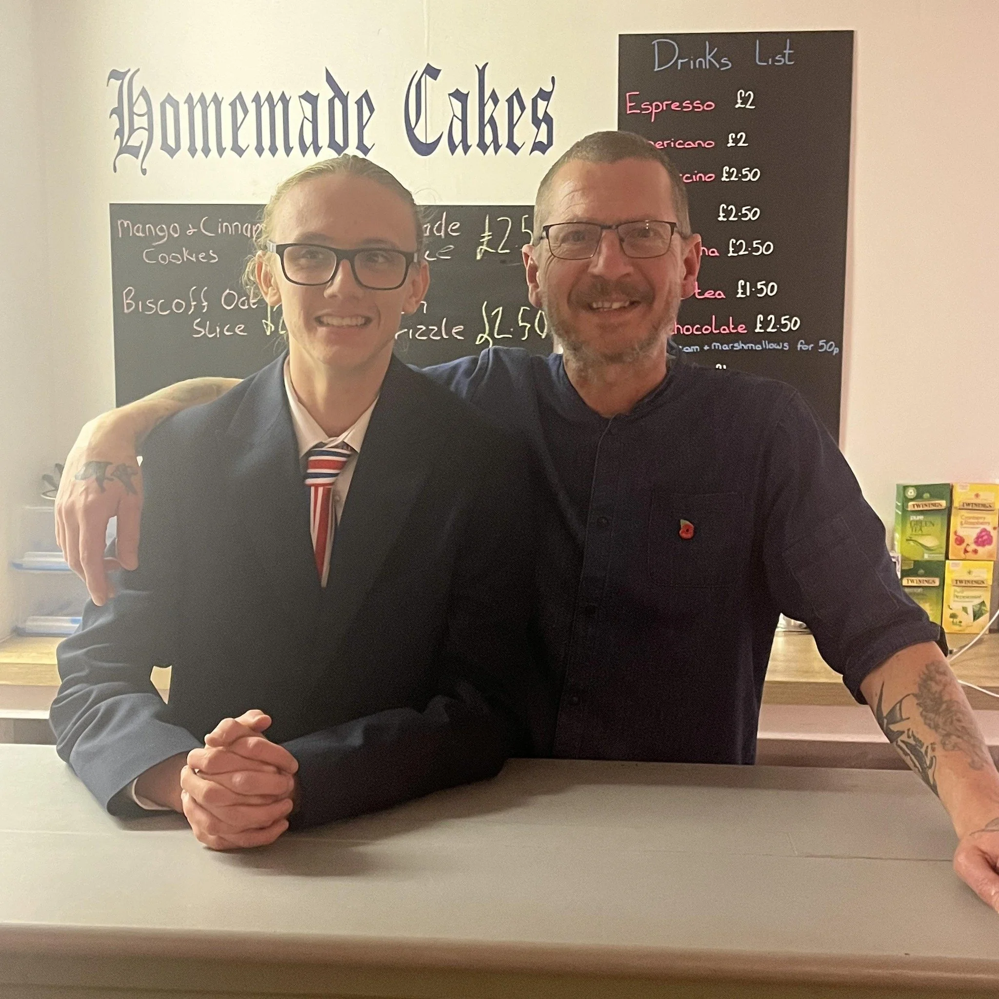 Two smiling men taking a selfie in front of a menu board. One man has glasses and tattoos, wearing a dark blazer with a striped tie. The other man has glasses and a beard. The menu board behind them lists drinks and prices, and there is a sign for homemade cakes.