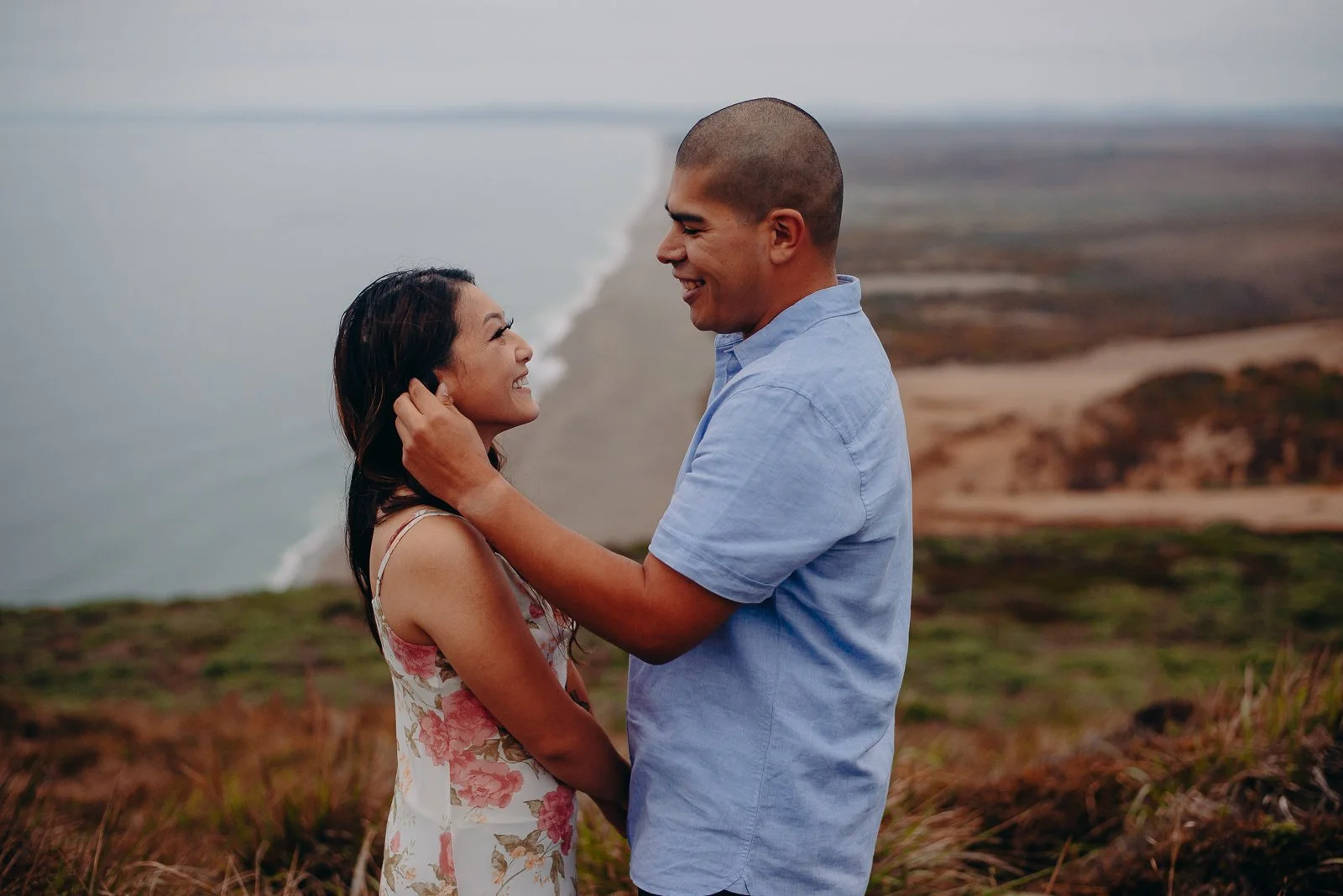 A smiling couple standing close together outdoors on a hilltop overlooking a coastline and ocean, facing each other, embracing, with the woman touching the man's face.
