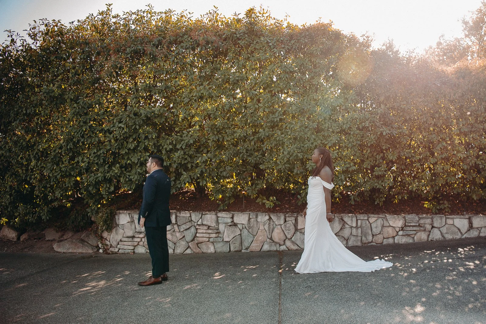 A bride and groom standing apart on a paved street with a stone wall and large green shrubbery behind them, during sunset.