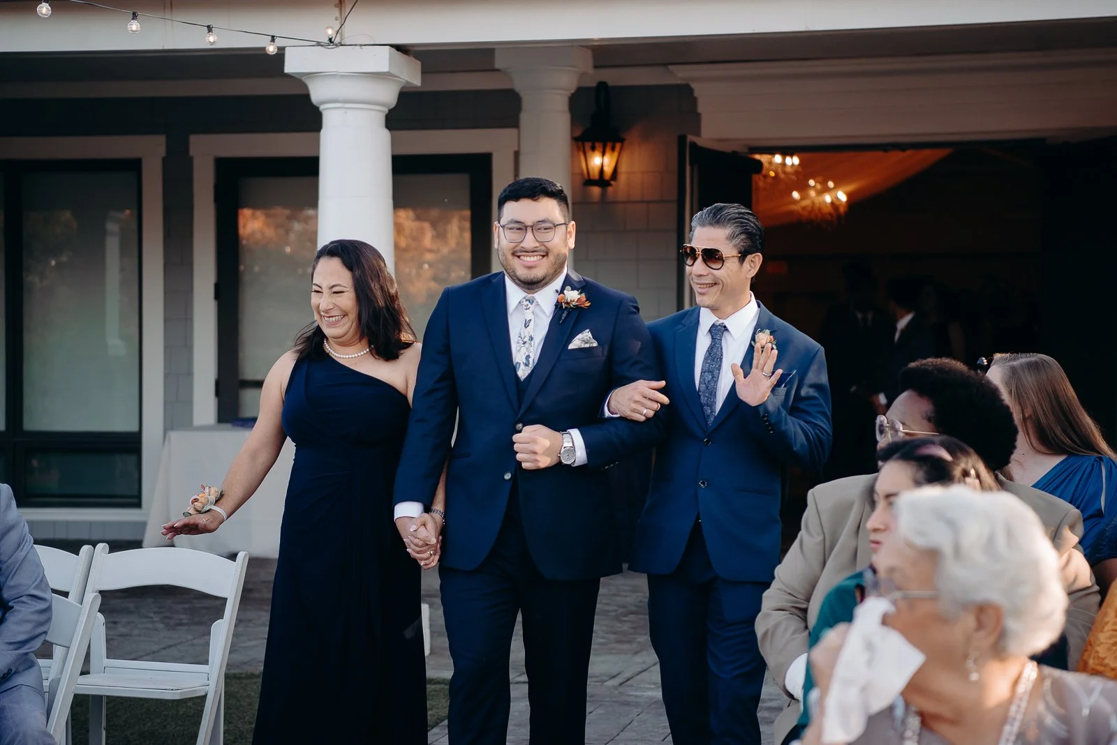A man in a blue suit and glasses holding hands with a woman in a blue dress, smiling. Another man in a blue suit and sunglasses is smiling and waving. Several seated guests are visible, some wearing glasses and colorful clothing, at an outdoor event.