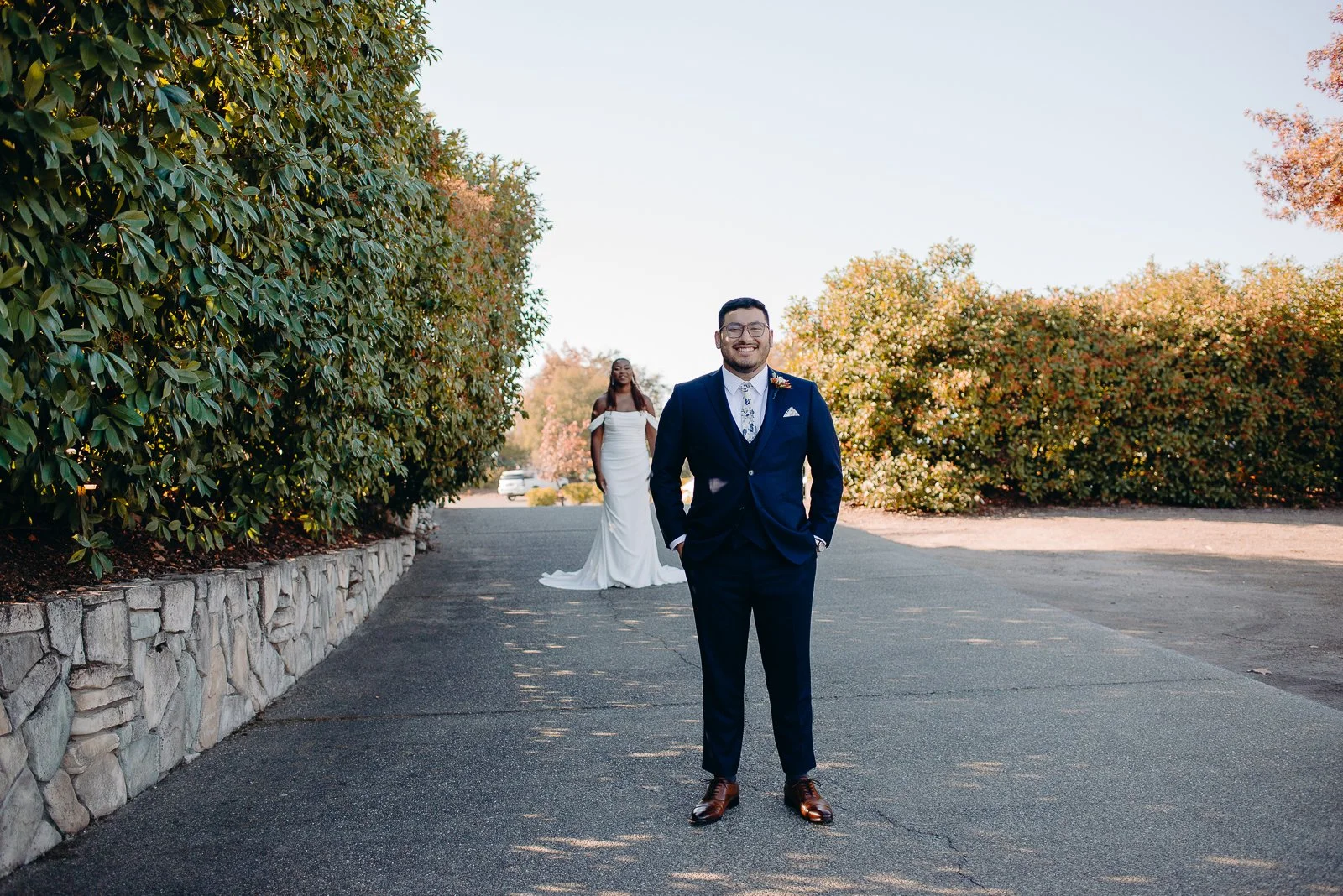 A happy groom in a navy blue suit and glasses stands with his hands in his pockets in the foreground, while a smiling bride in a white wedding dress stands in the background on a paved pathway surrounded by greenery and trees with autumn leaves.