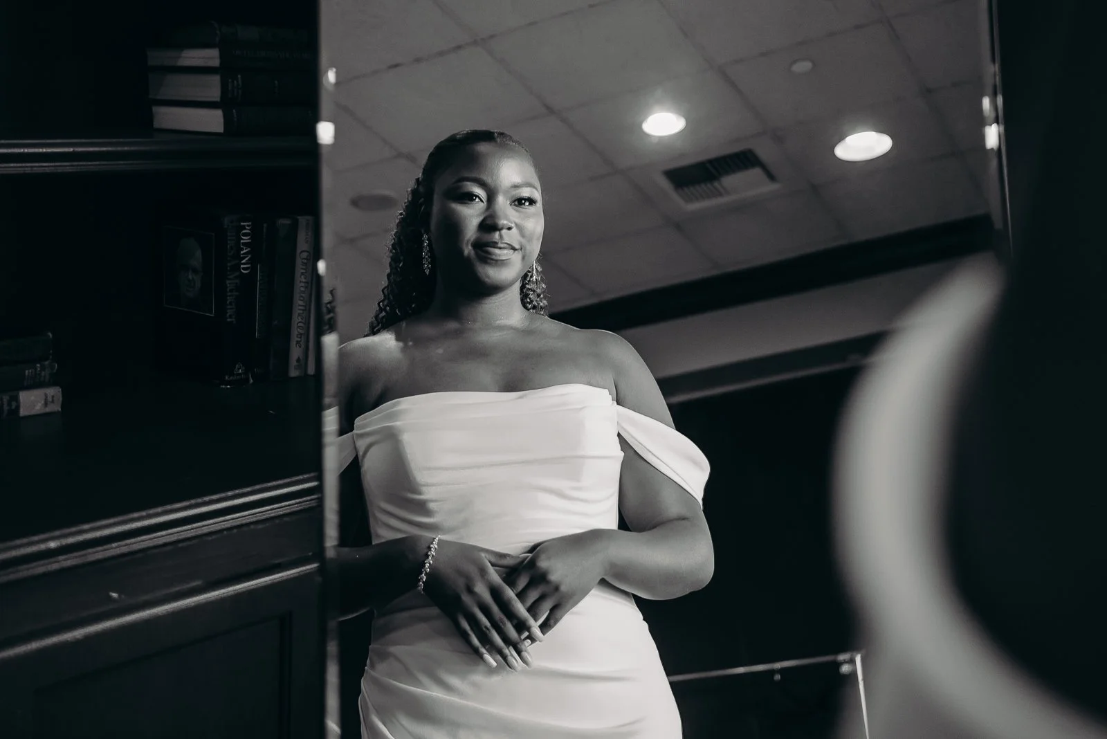 A woman in a strapless white dress standing near bookshelves in what appears to be a library or bookstore, with a ceiling light overhead and a window or reflection on her left.