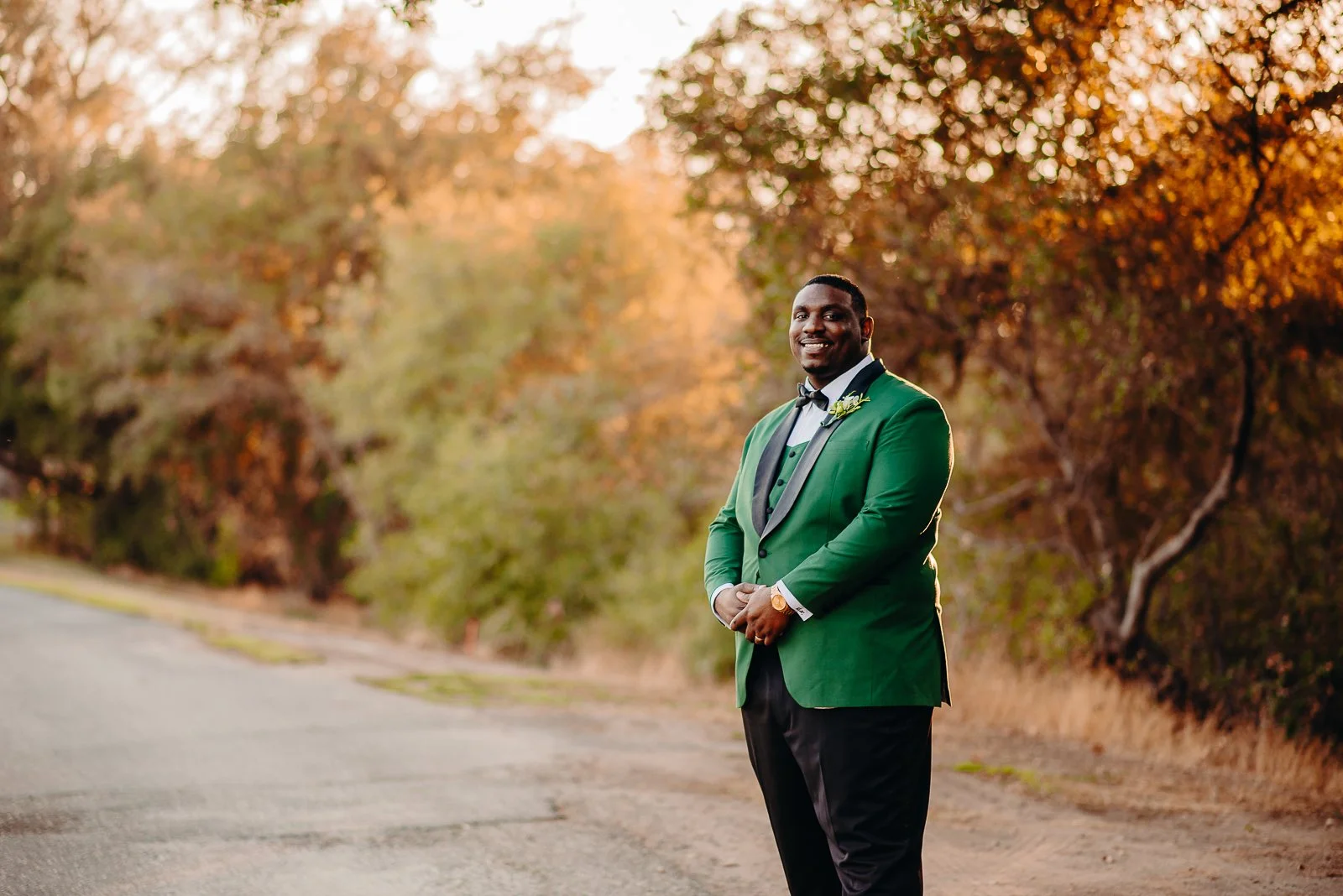 A man in a green tuxedo jacket and black pants stands outdoors on a paved road, smiling, with autumn trees in the background during sunset.