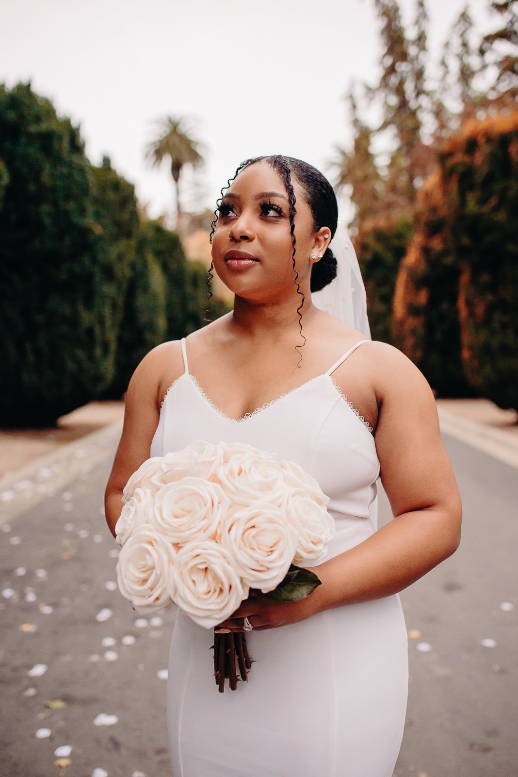A bride in a white dress holding a bouquet of beige roses, with trees and a road in the background.