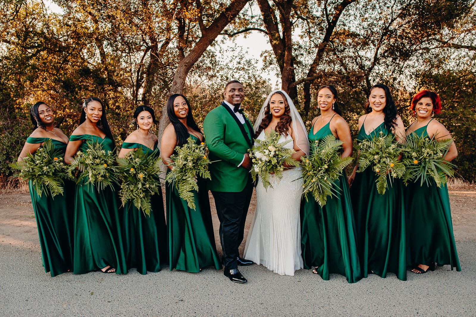 A wedding party standing outdoors in front of trees during sunset. The group consists of nine women in matching emerald green dresses and a man in a green tuxedo jacket with black pants, standing next to a bride in a white wedding gown with a bouquet