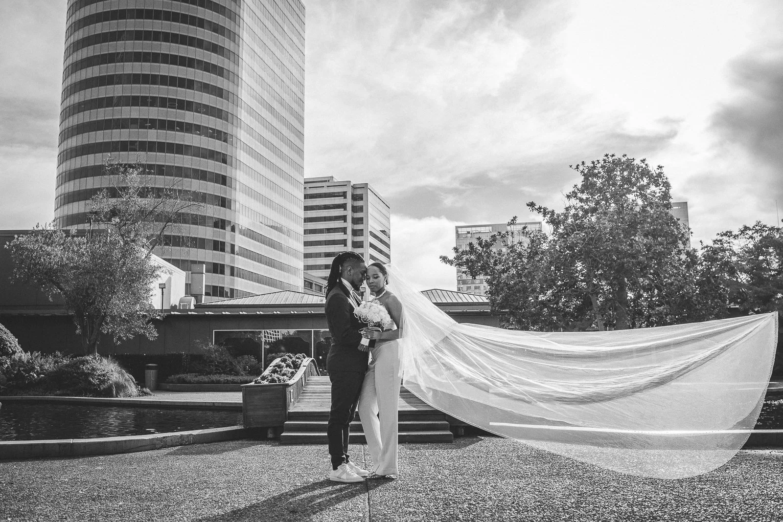 A bride and groom standing close together outdoors in front of a cityscape, with the bride holding a bouquet and her veil flowing behind her in the wind.
