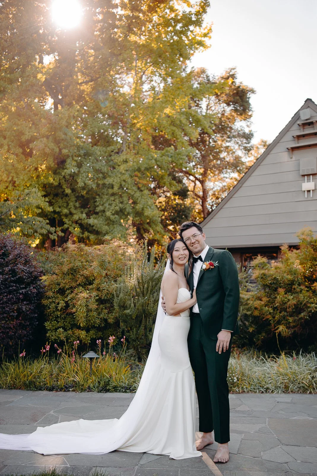 A newlywed couple smiling and hugging outdoors at sunset. The bride is in a white strapless wedding gown, and the groom is in a black tuxedo with a bow tie. The background features trees, bushes, and a house with a steep roof, bathed in warm sunlight