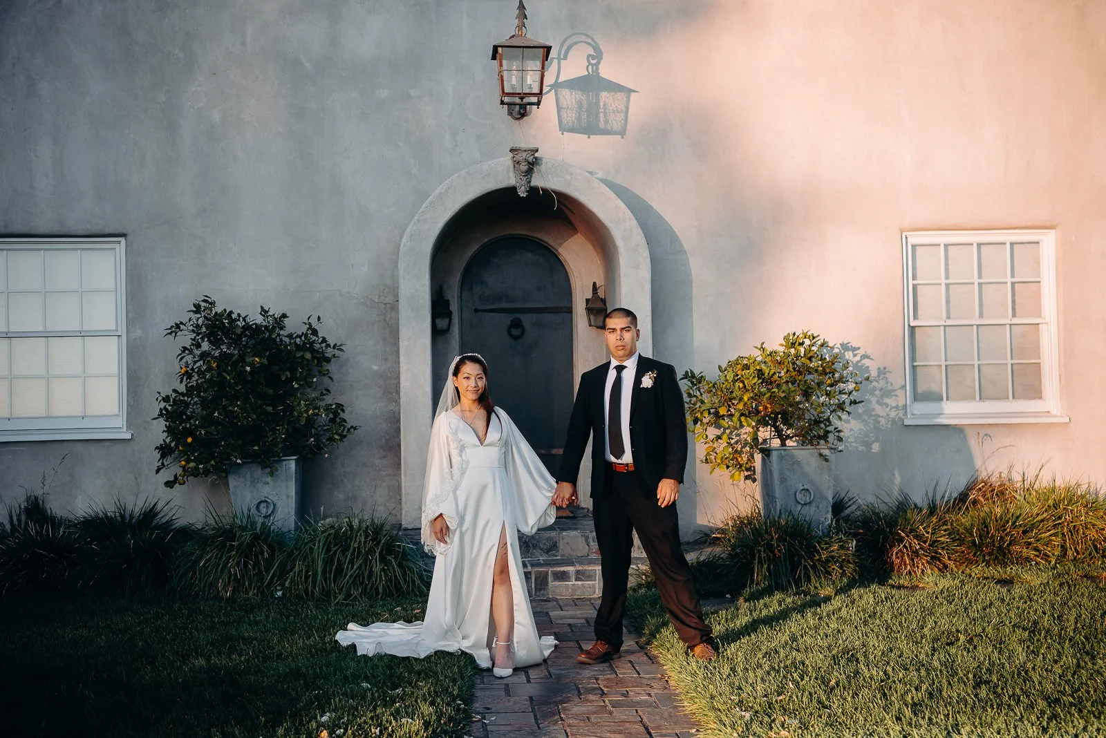 A newlywed couple is standing on a brick pathway in front of a white house with plants and grass. The bride is wearing a white wedding gown with a slit, veil, and is holding hands with the groom, who is dressed in a black suit and tie. They are holdi