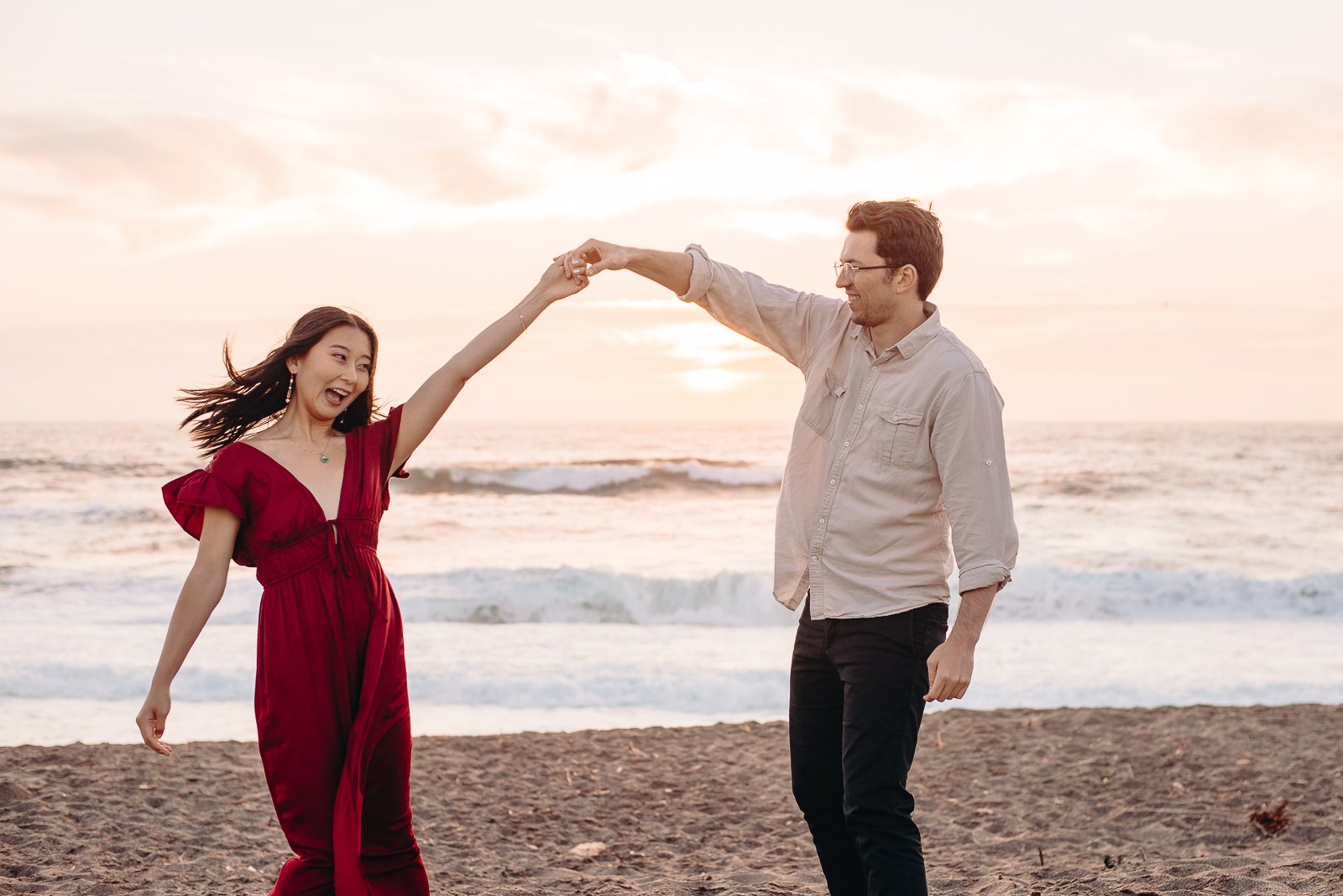 A couple on the beach at sunset, with the woman in a red dress and the man in a light-colored shirt, holding hands and smiling.