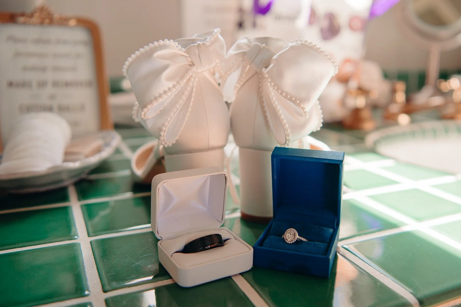 Wedding shoes, a wedding ring in a white box, and an engagement ring in a blue box on green tiled surface.