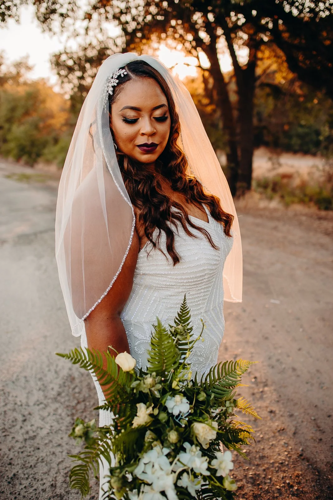 A bride in a white wedding dress holding a bouquet of white flowers and greenery, standing outdoors during sunset with trees in the background.