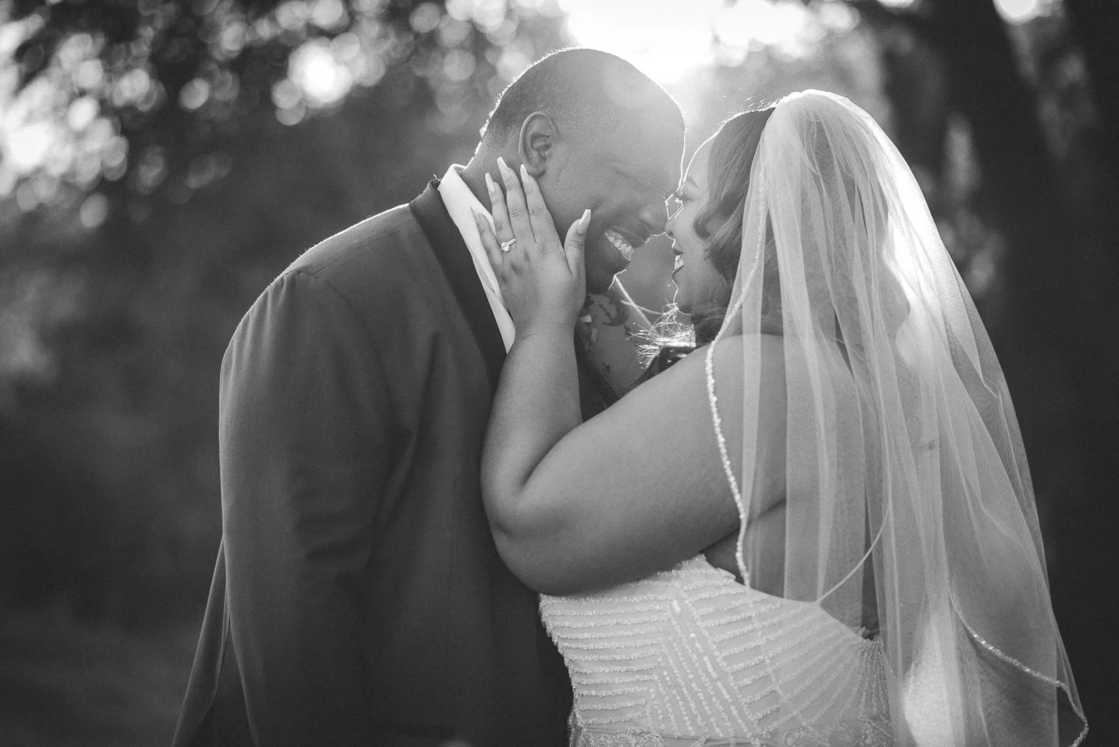 A couple on their wedding day, the groom and bride are facing each other, smiling, with the bride's hands on the groom's face. The bride is wearing a veil and a wedding dress, and the groom is in a suit. The scene is backlit by the sun, creating a ro