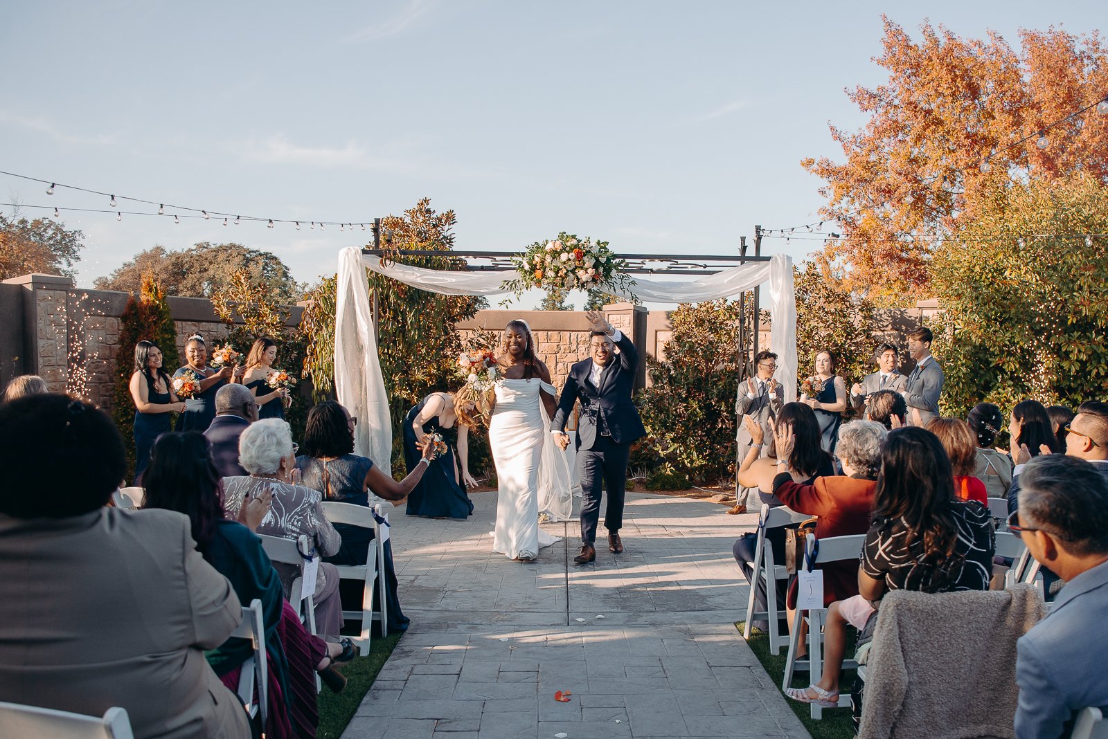 A wedding ceremony outdoors with the bride and groom holding hands, walking down the aisle, surrounded by friends and family. The ceremony area is decorated with white drapery and a floral arrangement overhead, with trees and string lights in the bac