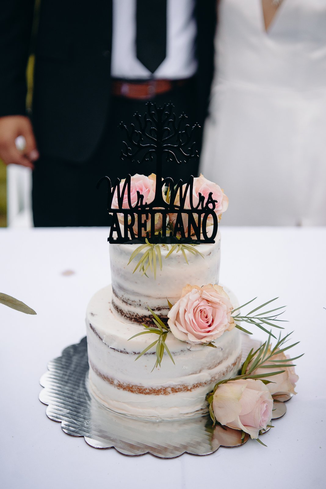 Two-tiered wedding cake with pink roses and greenery, topped with a black 'Mr & Mrs Arellano' cake topper, on a silver cake board, with a blurred couple in wedding attire in the background.