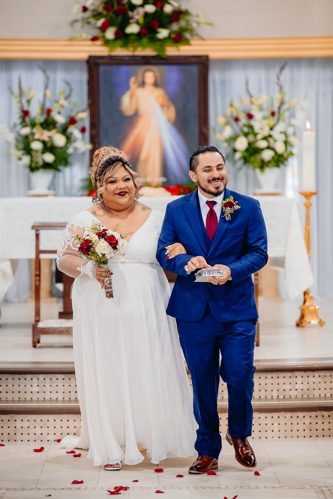 A bride and groom walk down the aisle in a church, smiling. The bride is dressed in a white gown with lace sleeves, holding a bouquet of red and white flowers. The groom is wearing a blue suit with a red tie. The background features a religious paint