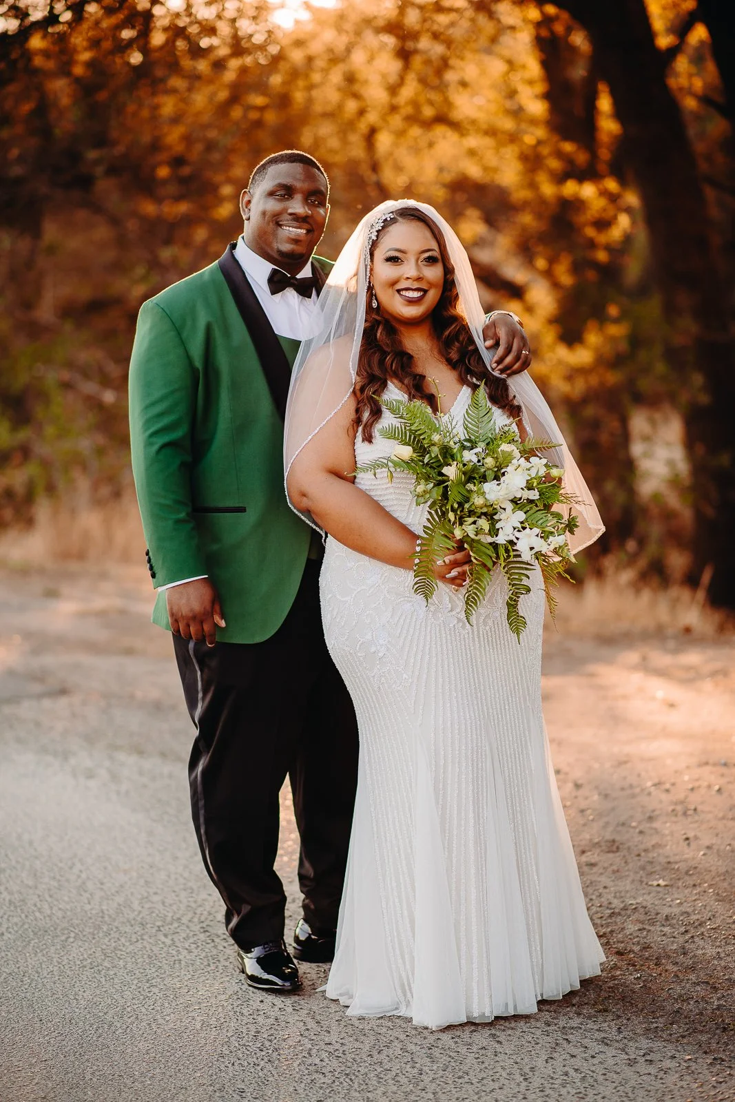 A bride and groom stand together outdoors during sunset. The bride is dressed in a white wedding gown with intricate patterns, holding a bouquet of white flowers and greenery. The groom is wearing a green tuxedo jacket with black lapels, a white shir