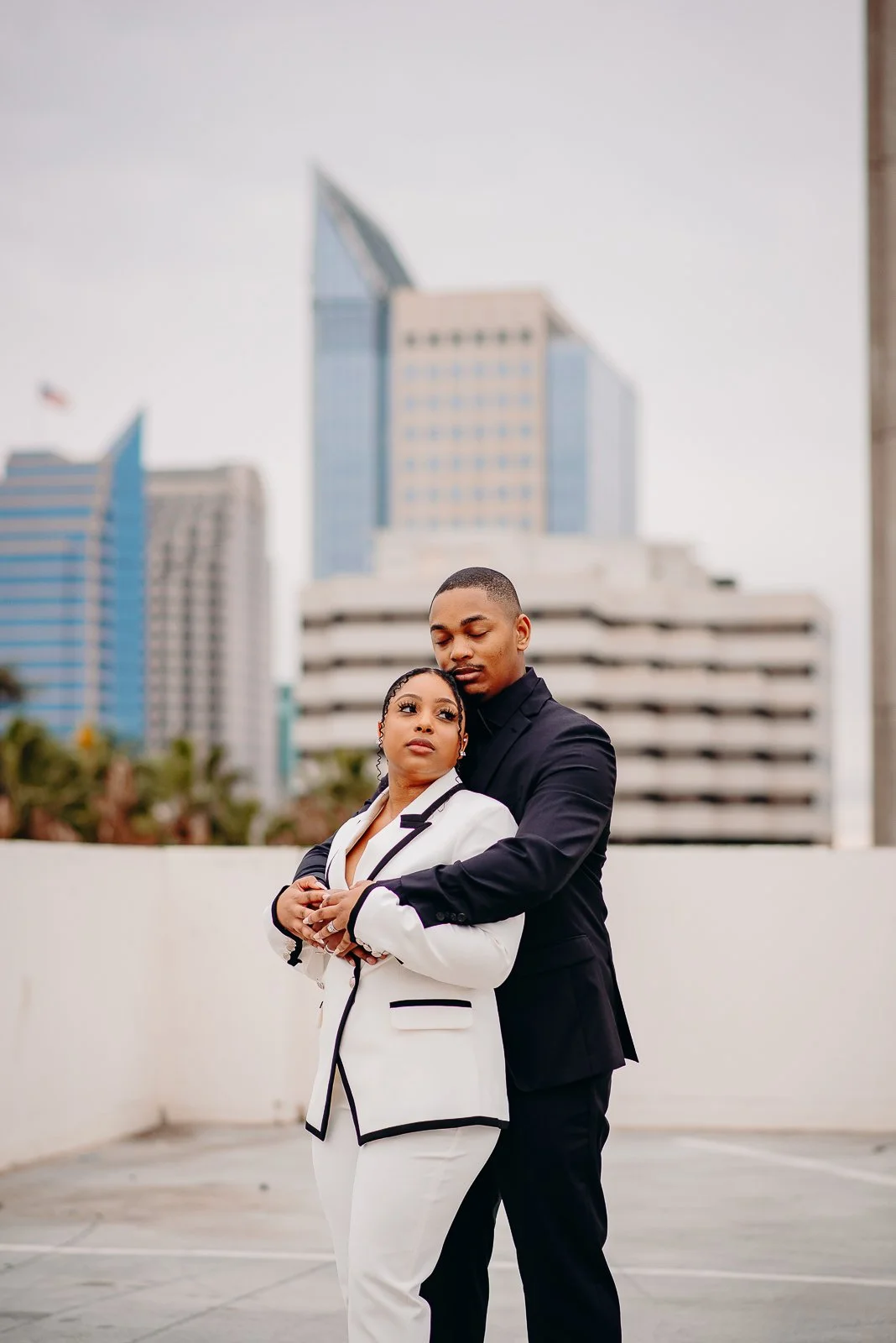 A couple in formal attire embracing on a rooftop with a city skyline in the background.