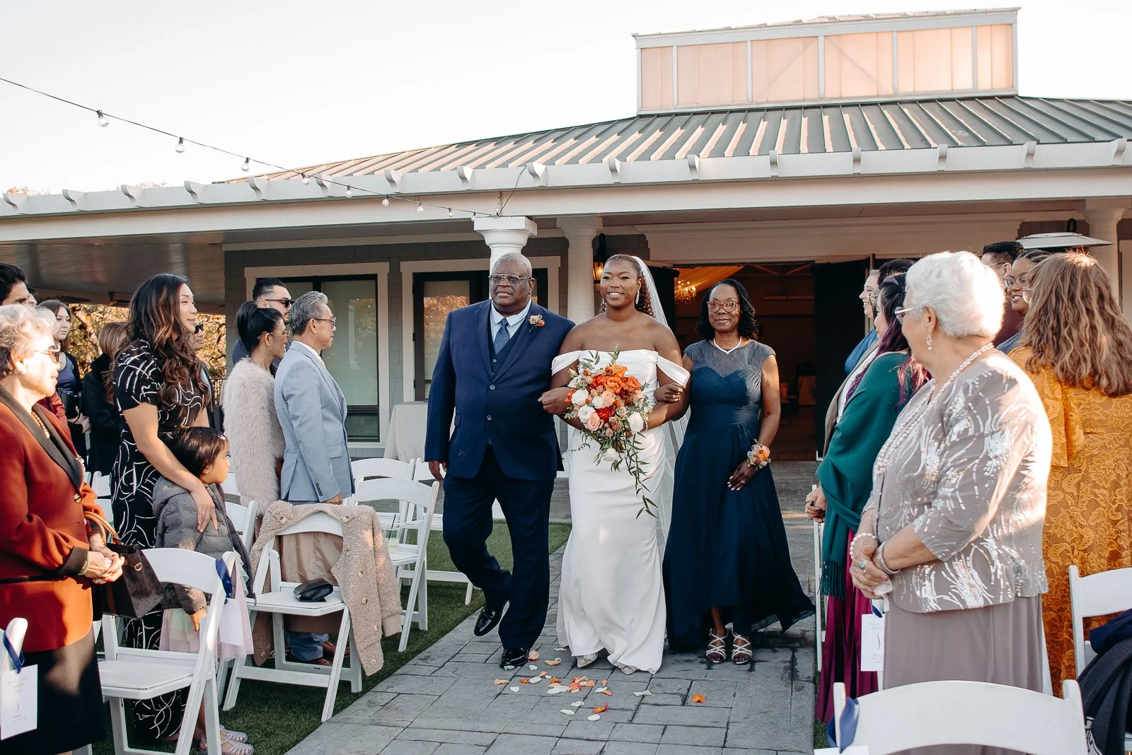 Bride walking down the aisle at a wedding reception, surrounded by guests, with a bouquet of flowers, outside venue, during daytime.