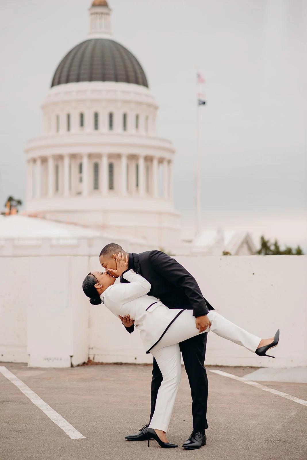 A couple in formal attire sharing a kiss on a parking garage rooftop with the California State Capitol building in the background.