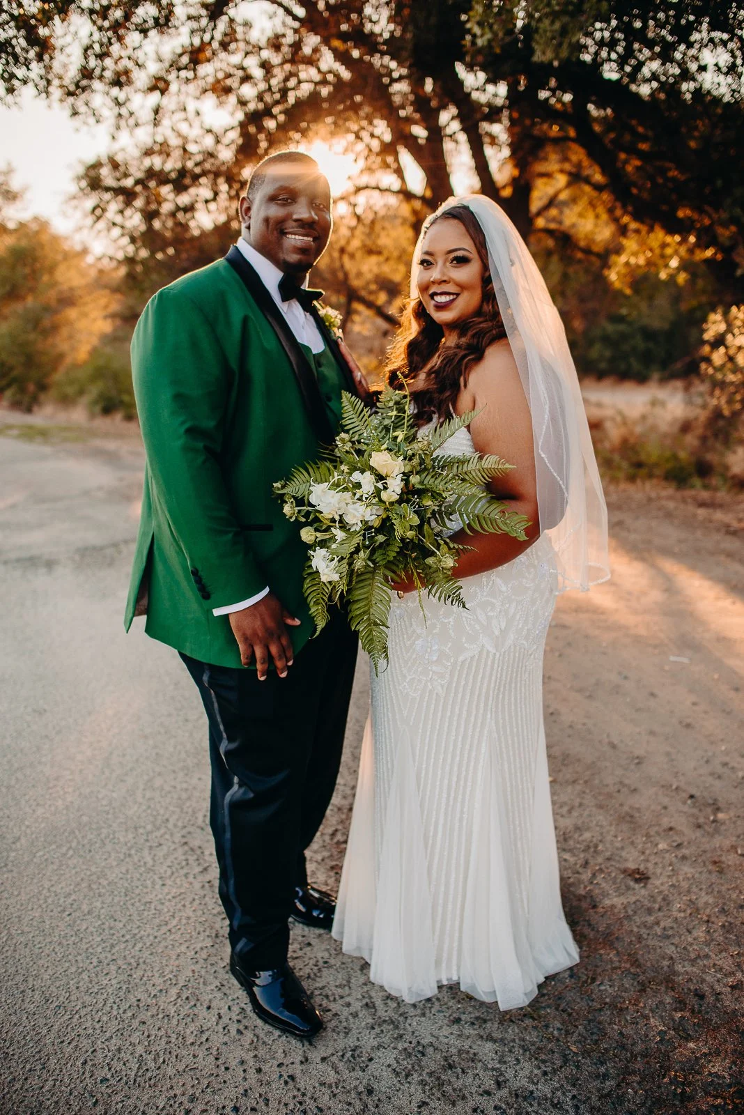 A newlywed couple is standing outdoors during sunset, with the bride holding a large bouquet of white flowers and greenery. The groom is dressed in a green tuxedo jacket and black tuxedo pants, and both are smiling at the camera.