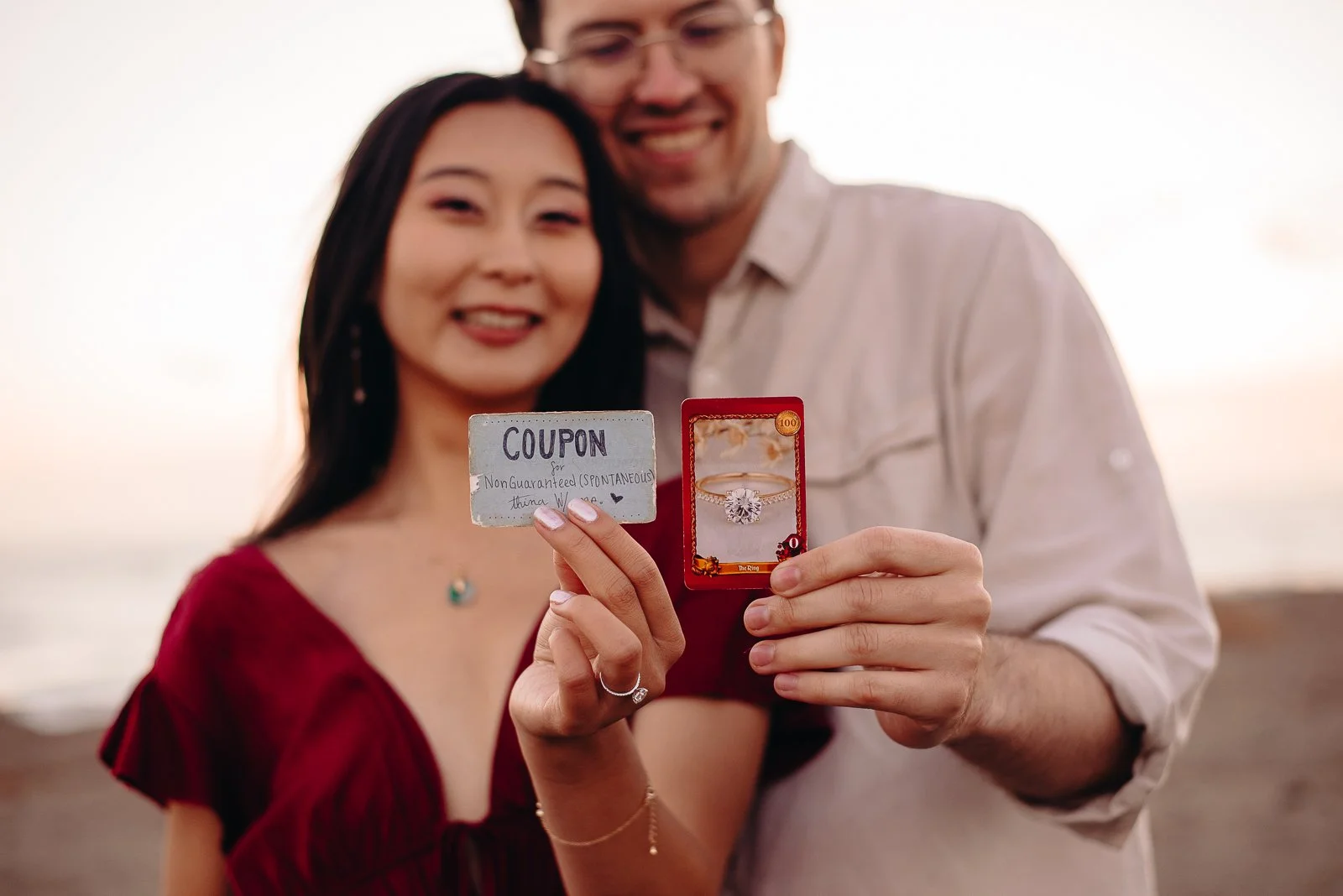 A happy couple posing outdoors at sunset, holding a box with a diamond engagement ring, the woman also holds a coupon note.