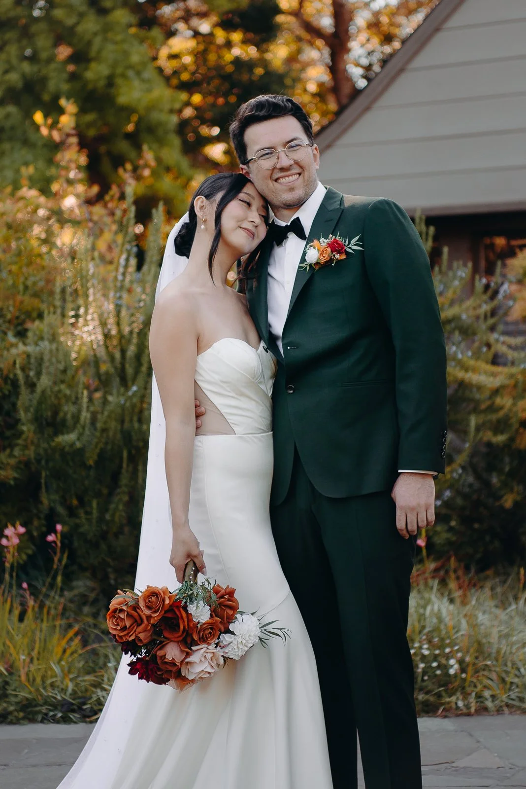 A bride and groom hugging outdoors during sunset, the bride holding a bouquet of orange, white, and blush flowers, and the groom wearing a dark green tuxedo with a boutonnière, surrounded by autumn foliage.