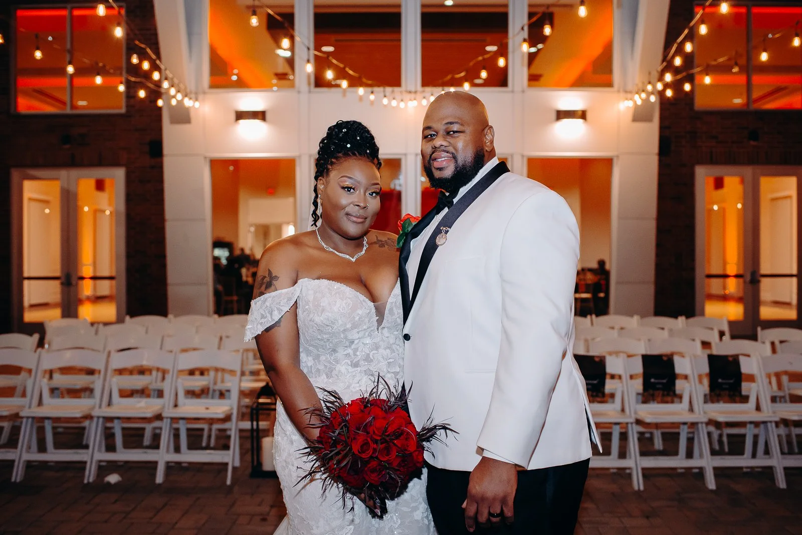 A bride and groom at their wedding reception, standing indoors in front of empty chairs and warm lighting, with the bride holding a bouquet of red roses and dark feathers.