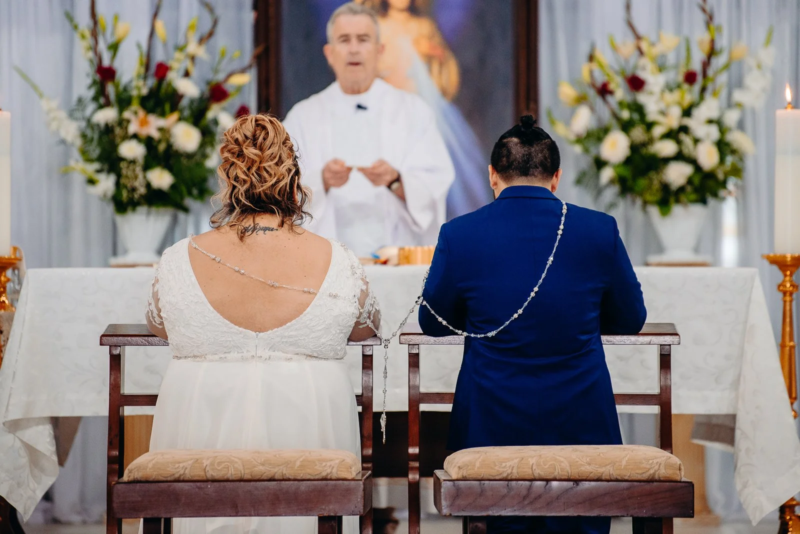 A couple kneeling during a wedding ceremony, with a priest officiating. The bride has curly hair and is wearing a white dress, while the groom has dark hair and is dressed in a blue suit. They are seated in front of an altar with floral arrangements 