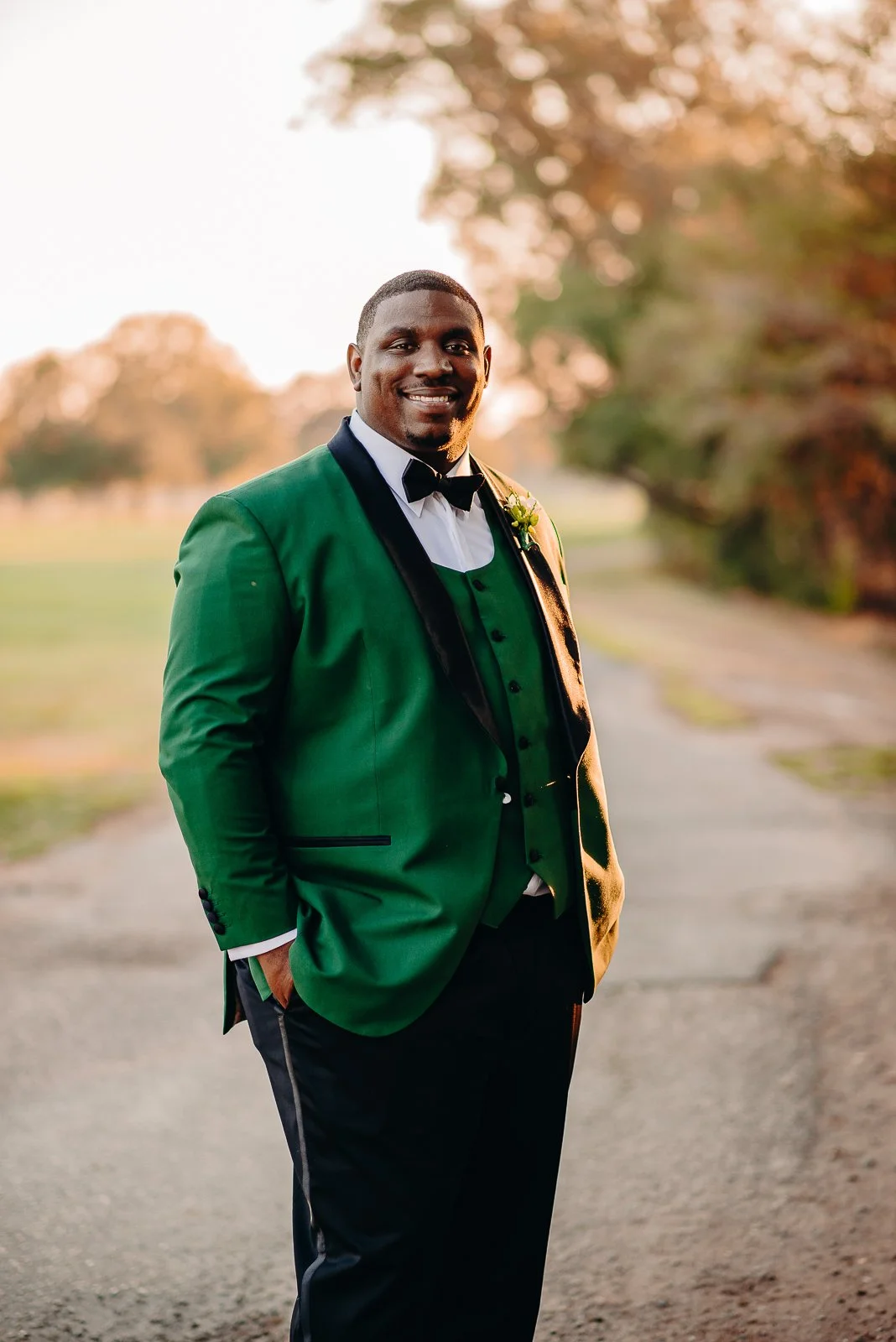 A man dressed in a green tuxedo jacket, white shirt, black bow tie, and black trousers standing outdoors on a pathway with trees in the background during sunset, smiling with hands in pockets.