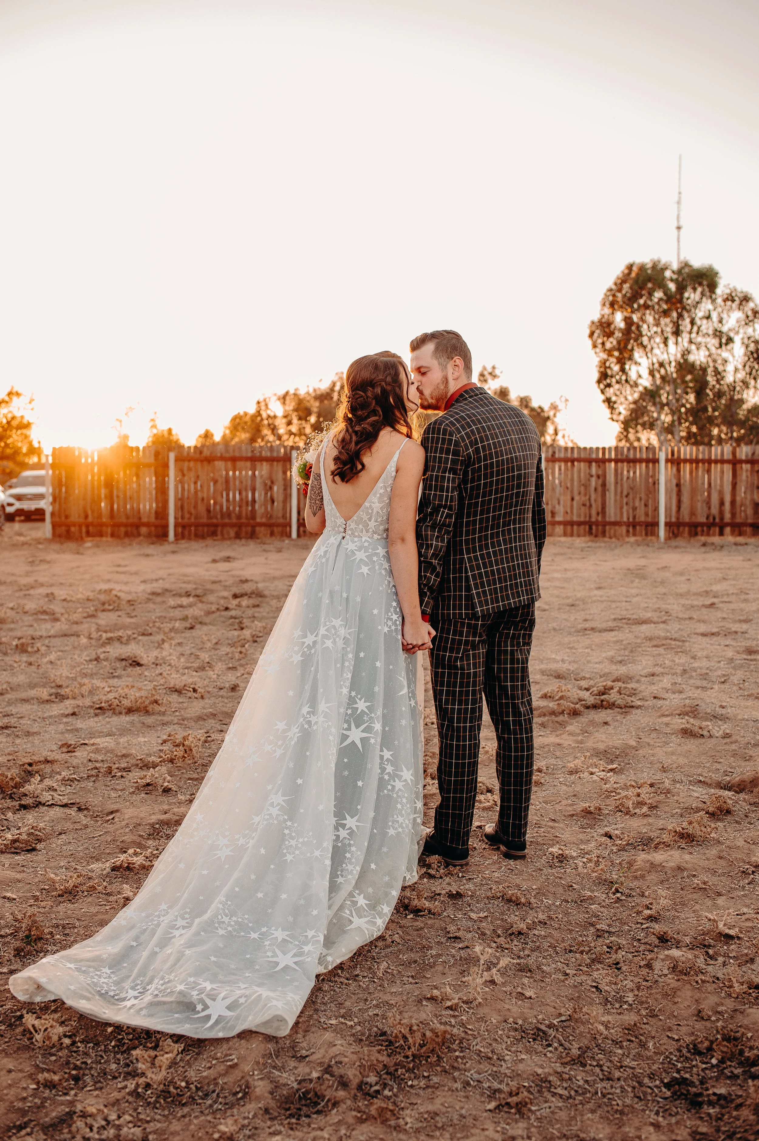 A bride and groom holding hands and about to kiss during sunset outdoors, with a wooden fence and trees in the background.