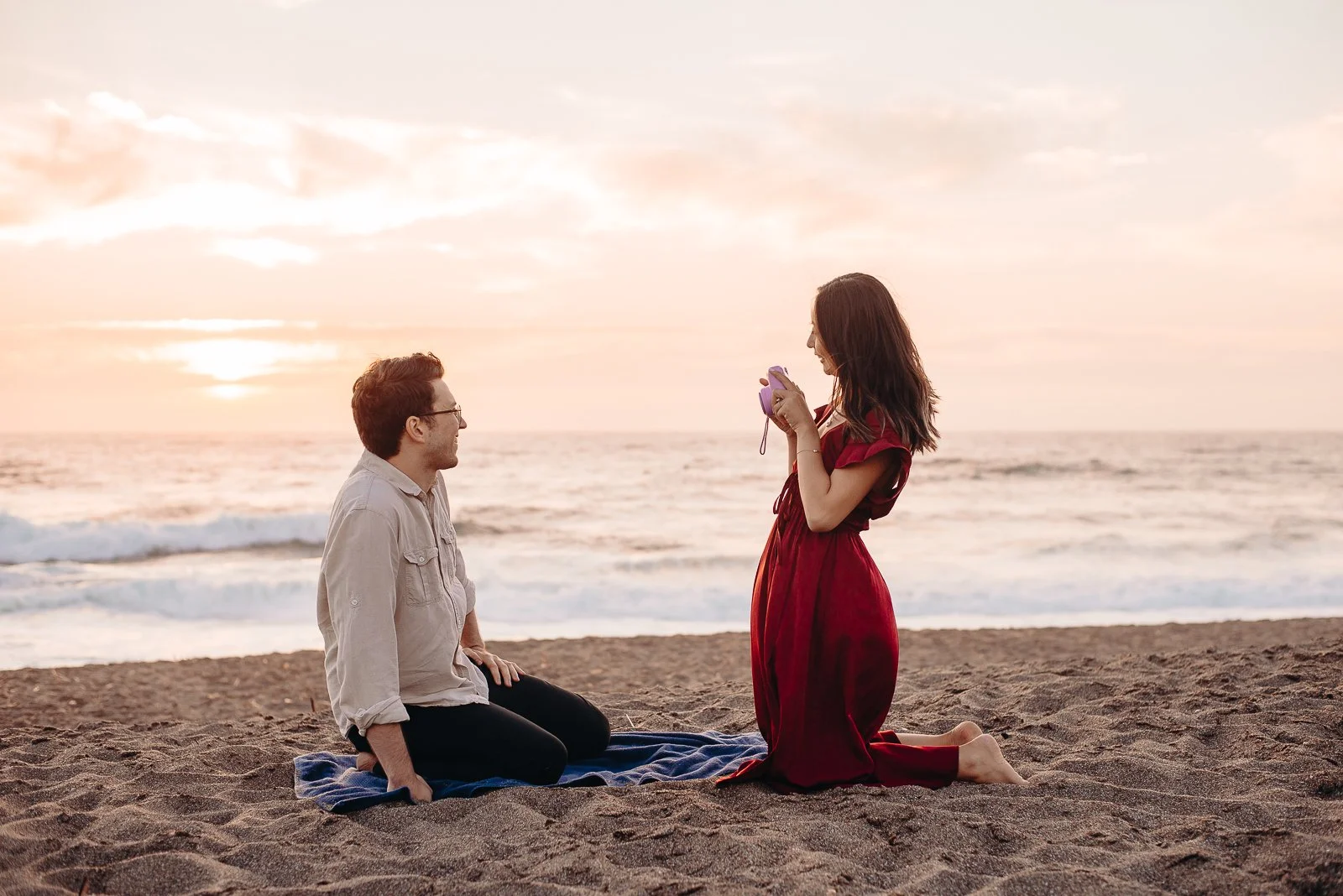A man kneeling on the sand at the beach, facing a woman who is kneeling on a blanket with a camera in her hands, during sunset.