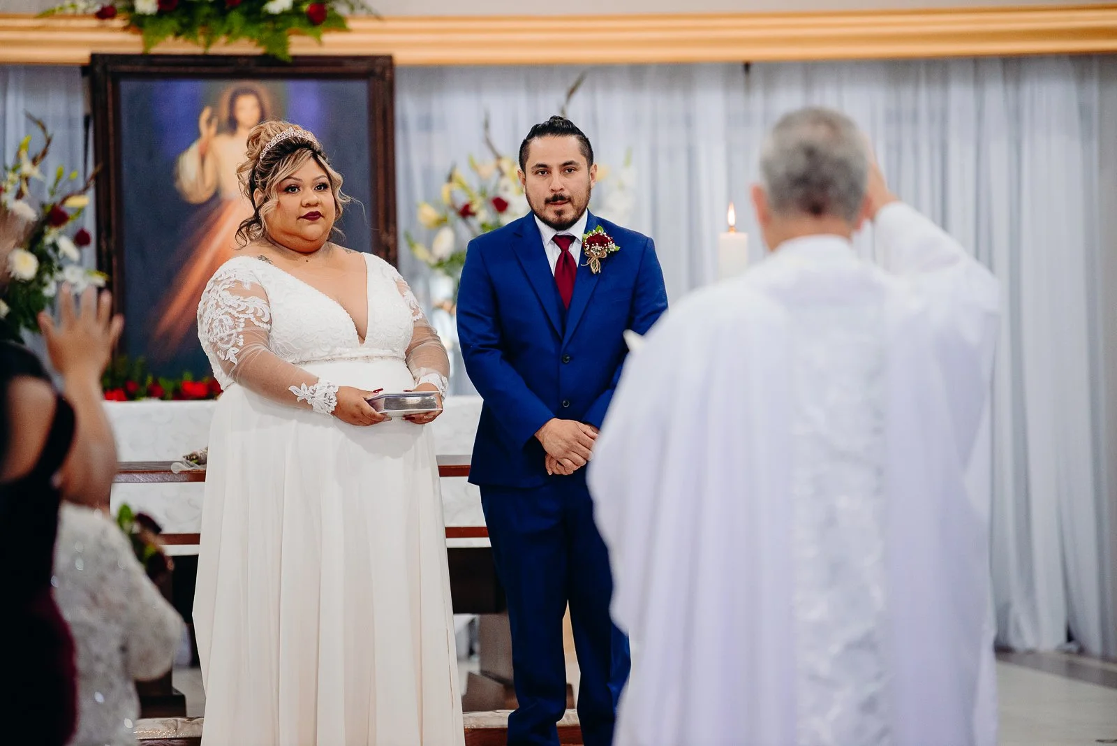 A couple standing at an altar during a wedding ceremony, with a priest in white robes conducting the service inside a church decorated with flowers and a religious painting in the background.