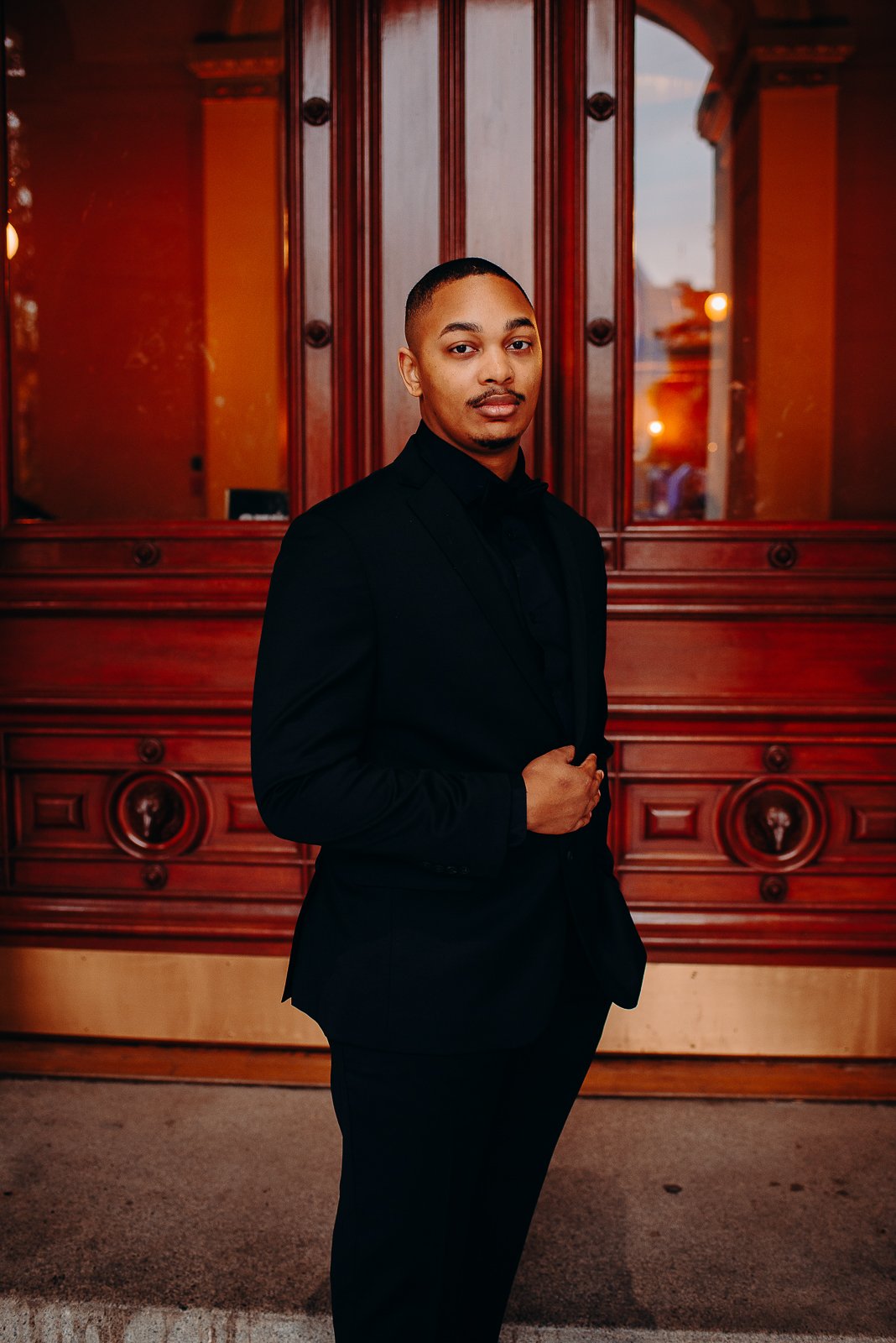A man in a black suit standing in front of a wooden door with ornate carvings, indoors, with lighting from the outside.