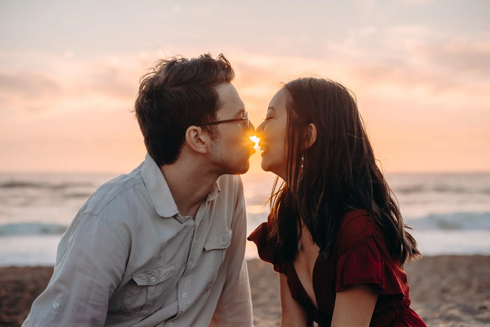A couple on the beach at sunset about to kiss with their noses touching and a sunset between their faces.
