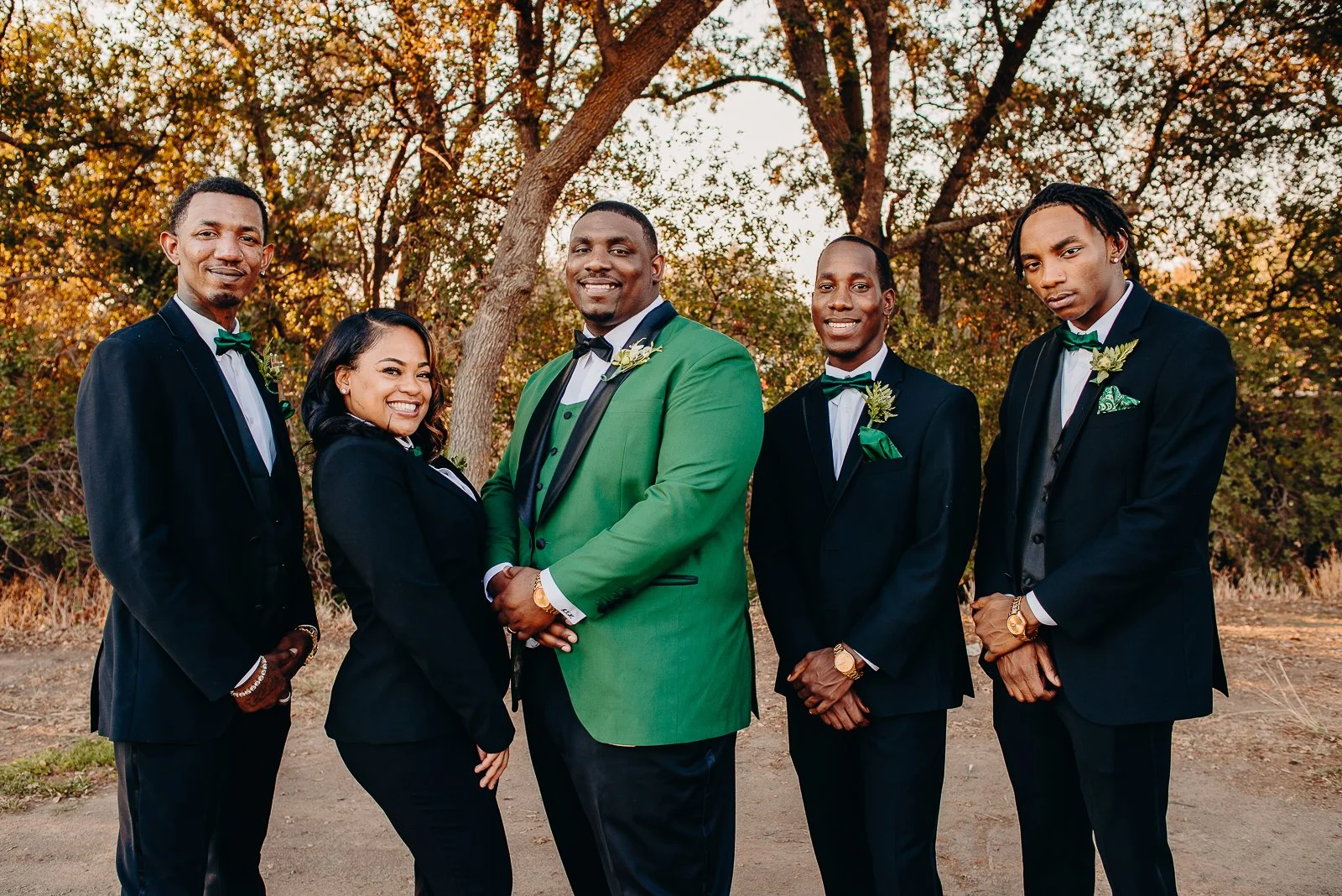 Group of six people dressed in formal wedding attire standing outdoors among trees, with the bride and groom in the center smiling, during sunset or late afternoon.