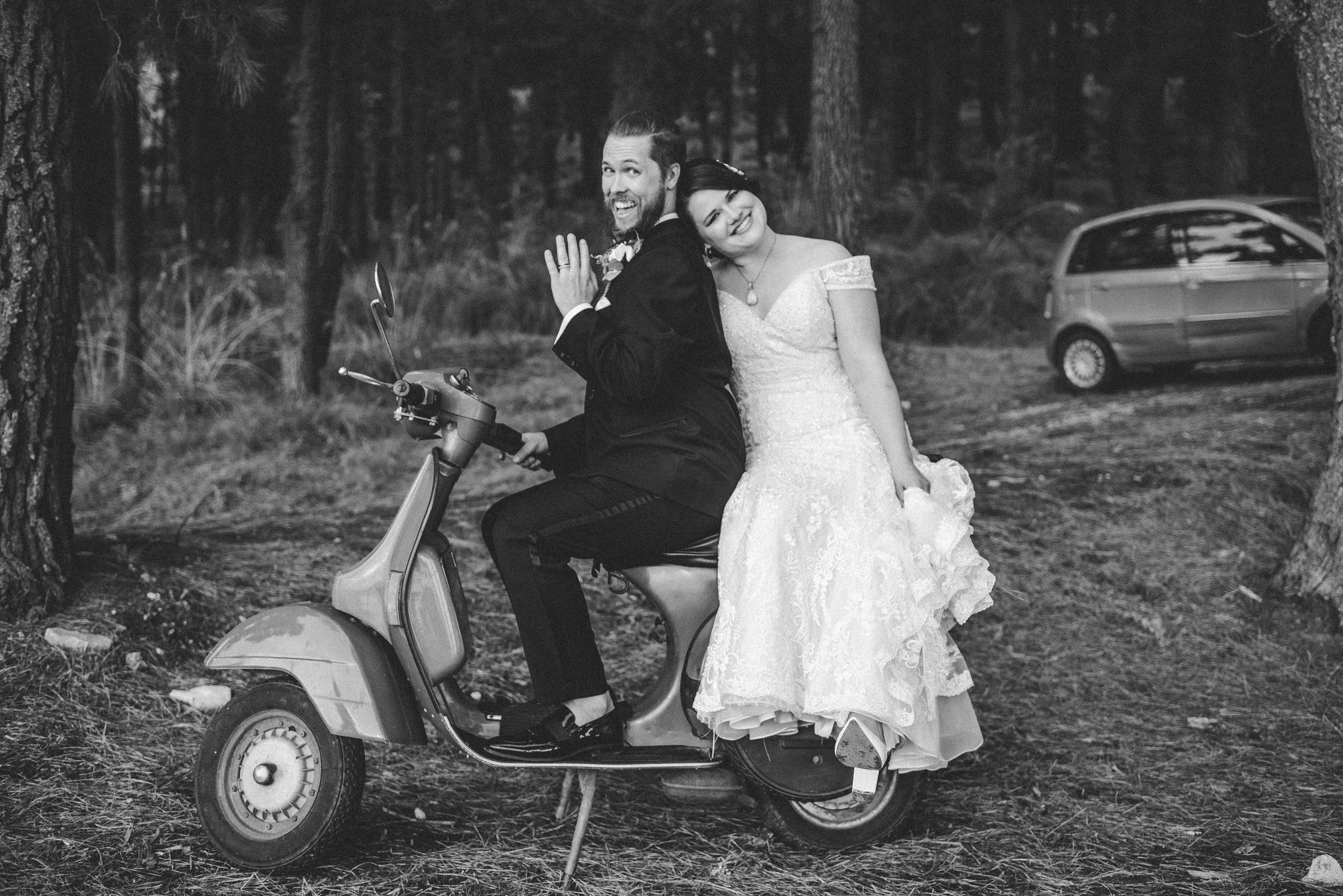 A wedding couple on a scooter in a forest, with the bride sitting behind the groom. The groom is wearing a suit and the bride is in a wedding dress. There's a car parked in the background to the right.