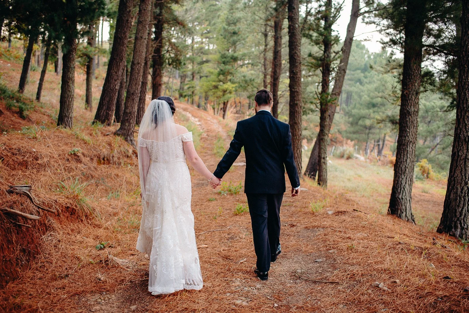 A bride and groom walking hand in hand through a forested trail.