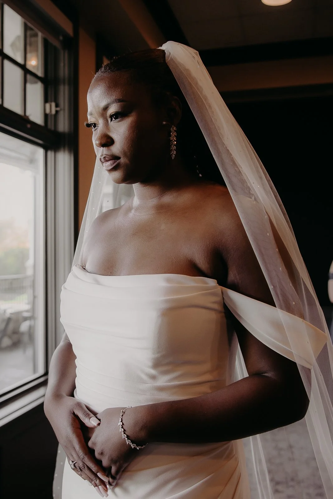 A bride in a strapless white wedding gown with a veil, standing indoors near a window, looking contemplative.