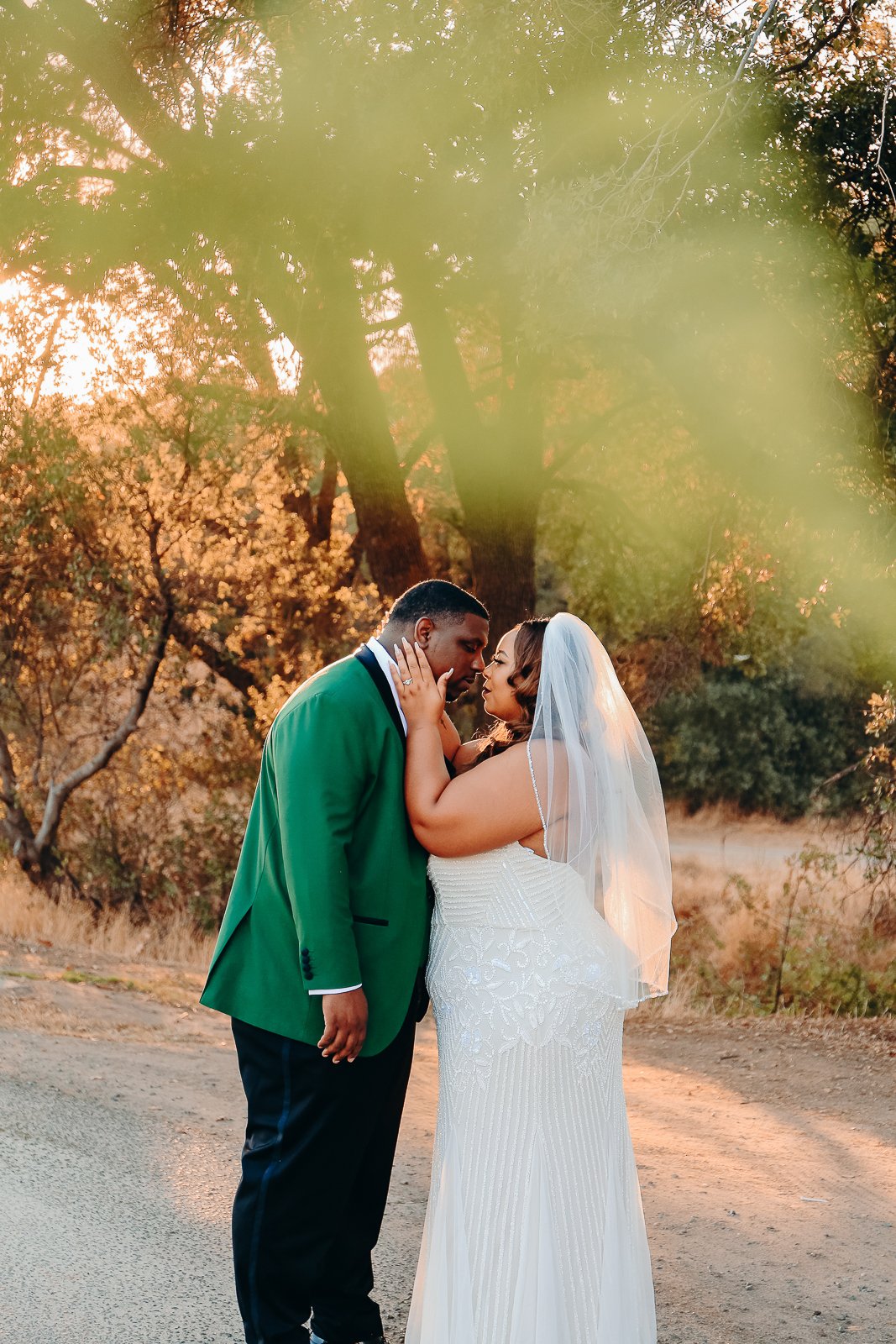 A bride and groom touch foreheads and hold each other's faces in an outdoor wedding photo during sunset, with trees and warm light in the background.