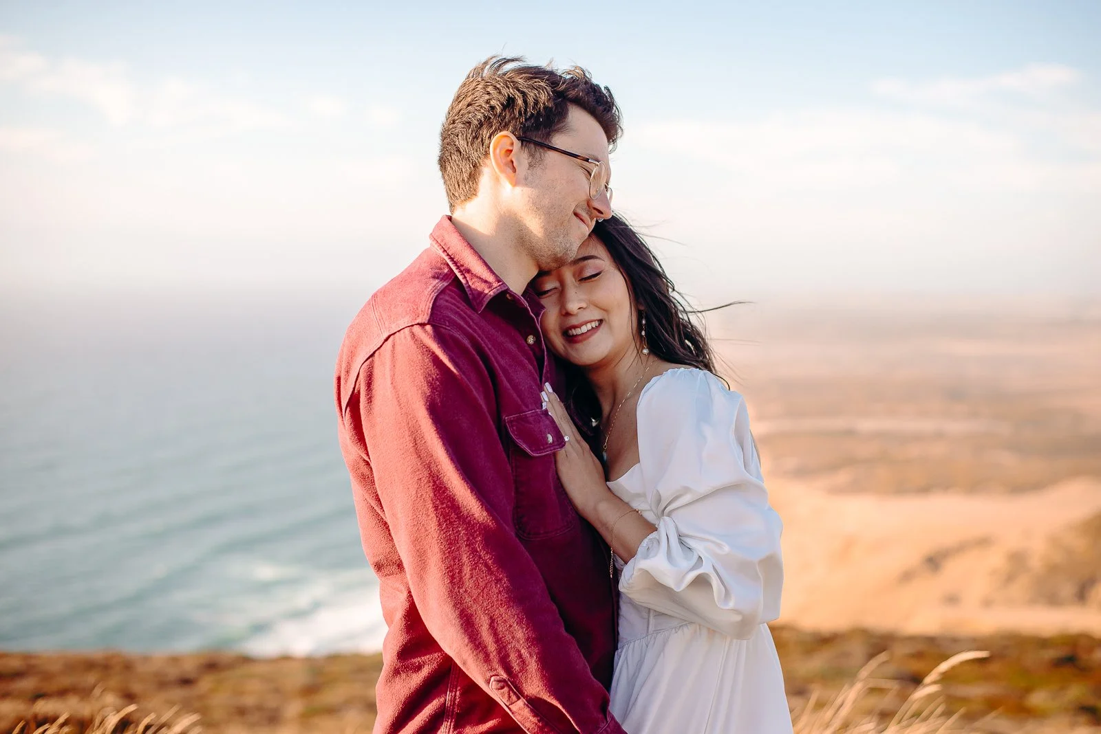 A couple embracing on a cliff overlooking the ocean, with the woman resting her head on the man's shoulder.