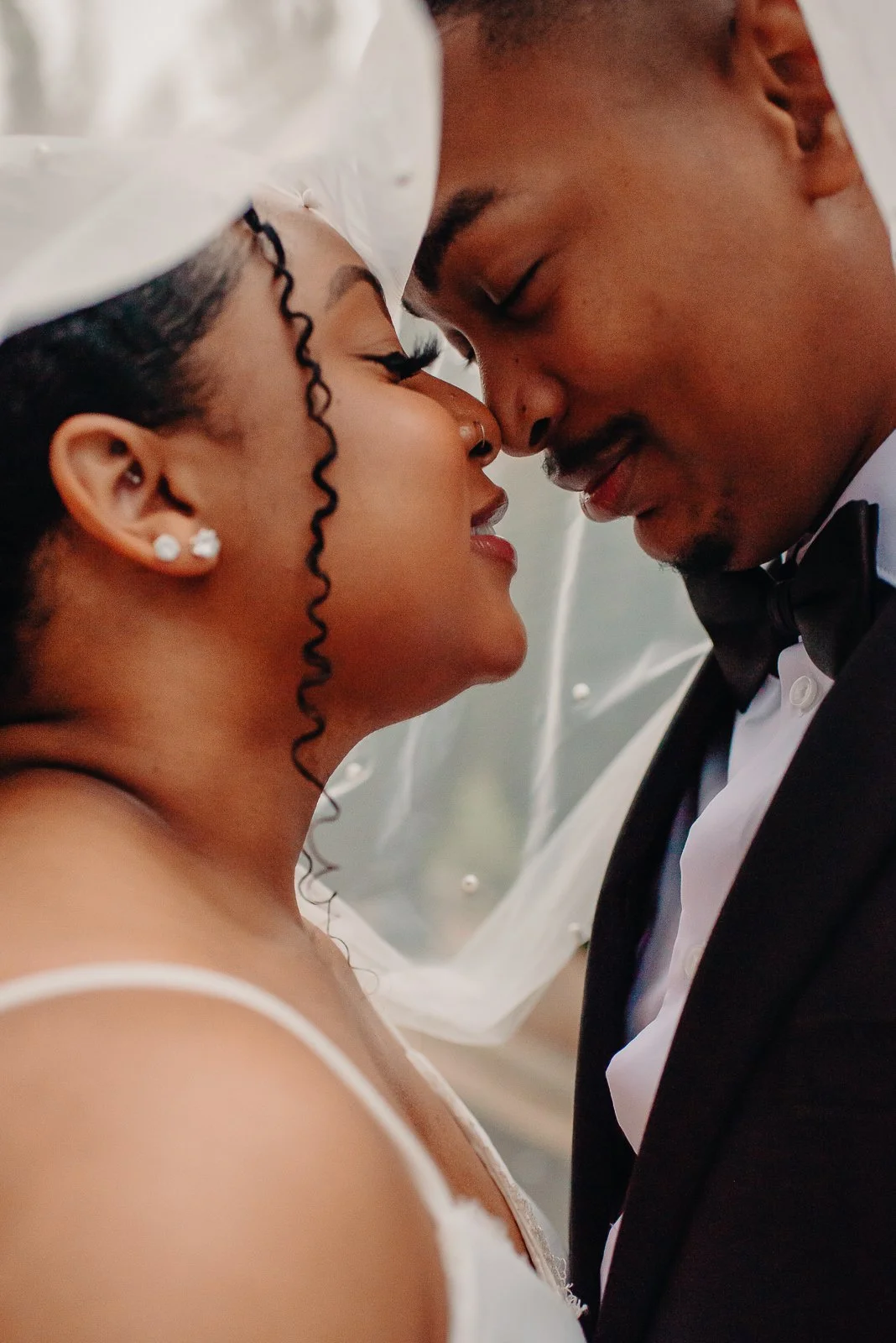 A bride and groom embrace with foreheads touching, eyes closed, in a wedding setting. The bride wears a veil, earrings, and a white dress. The groom wears a tuxedo and bow tie.