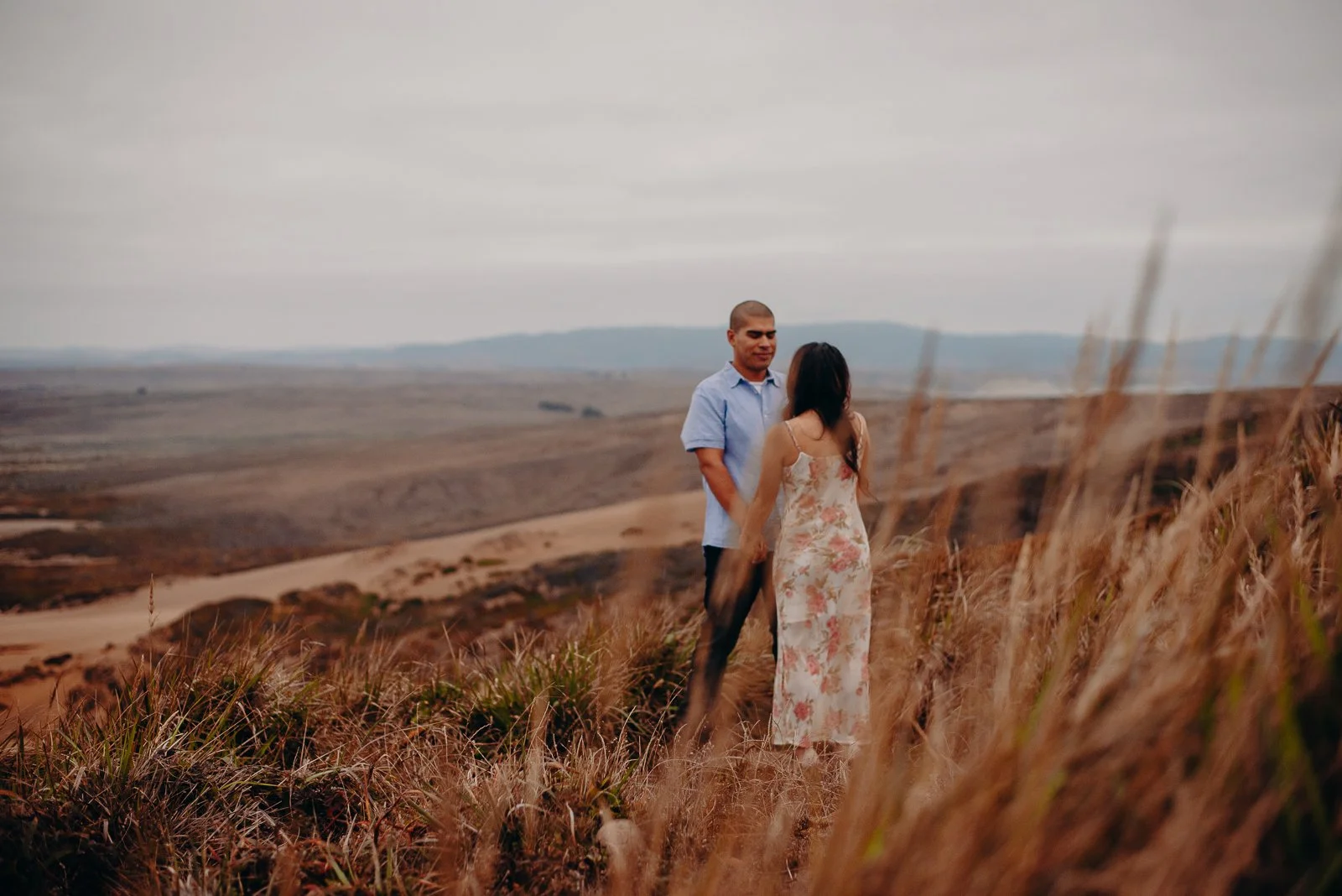 A couple holding hands on a grassy hillside with open landscape and mountains in the background under a cloudy sky.